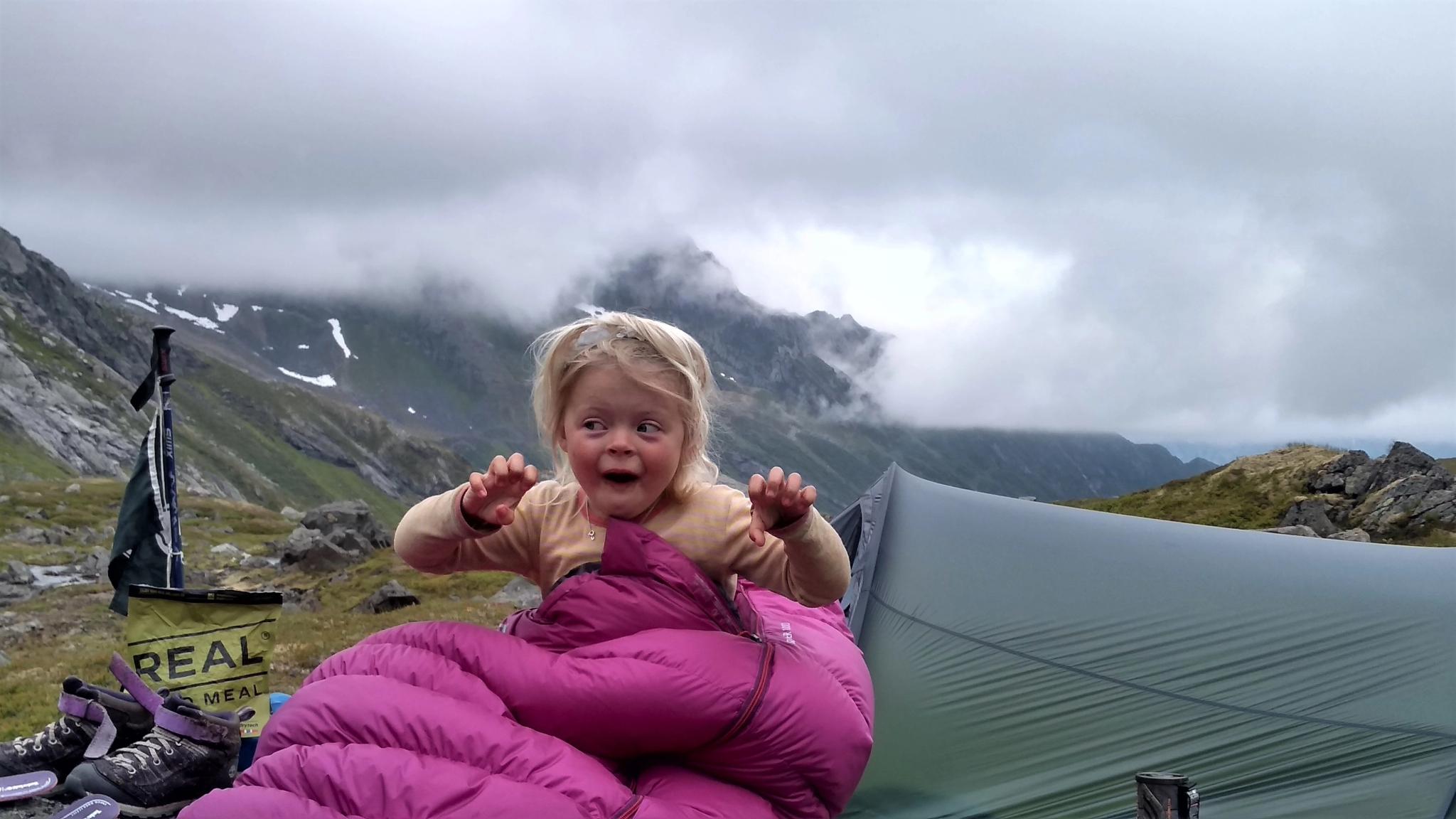 A young girl sitting in a sleeping bag imitating a troll outside a tent by the Litlekoppvatnet lake in Hjørundfjorden, Fjord Norway