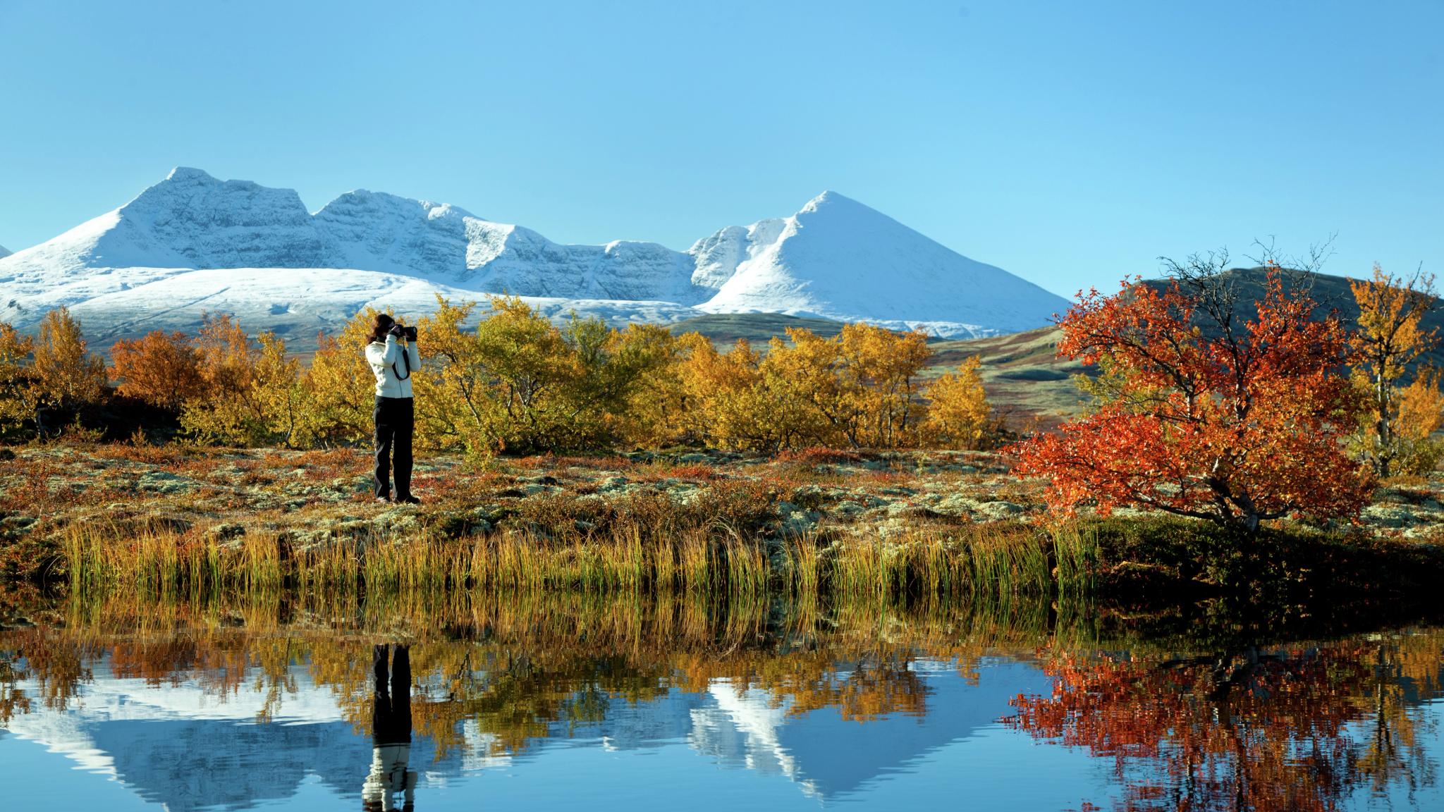 A woman taking photos of a lake and trees in autumn colours in the Rondane national park, Eastern Norway. Snowclad mountains in the background.