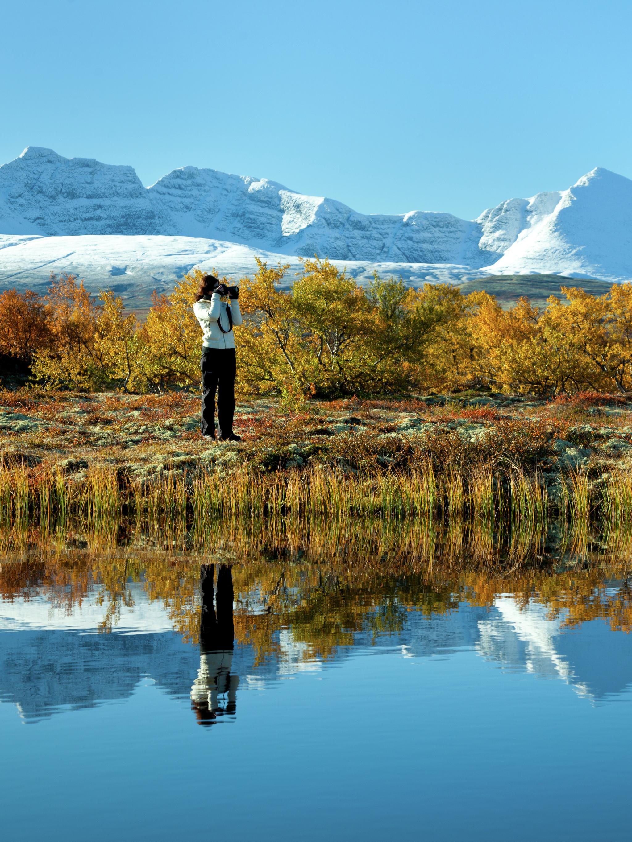 A woman taking photos of a lake and trees in autumn colours in the Rondane national park, Eastern Norway. Snowclad mountains in the background.