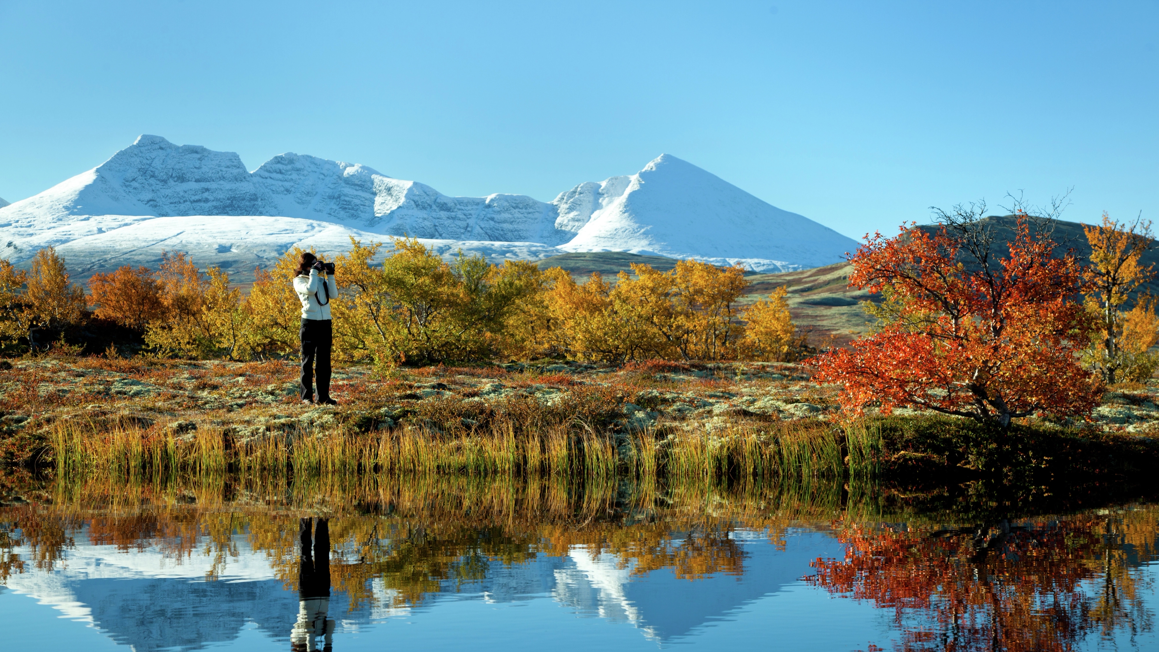 En kvinna tar höstbilder av en sjö och träd i Rondane nationalpark i Østlandet (östra Norge). I bakgrunden syns snöklädda fjäll.