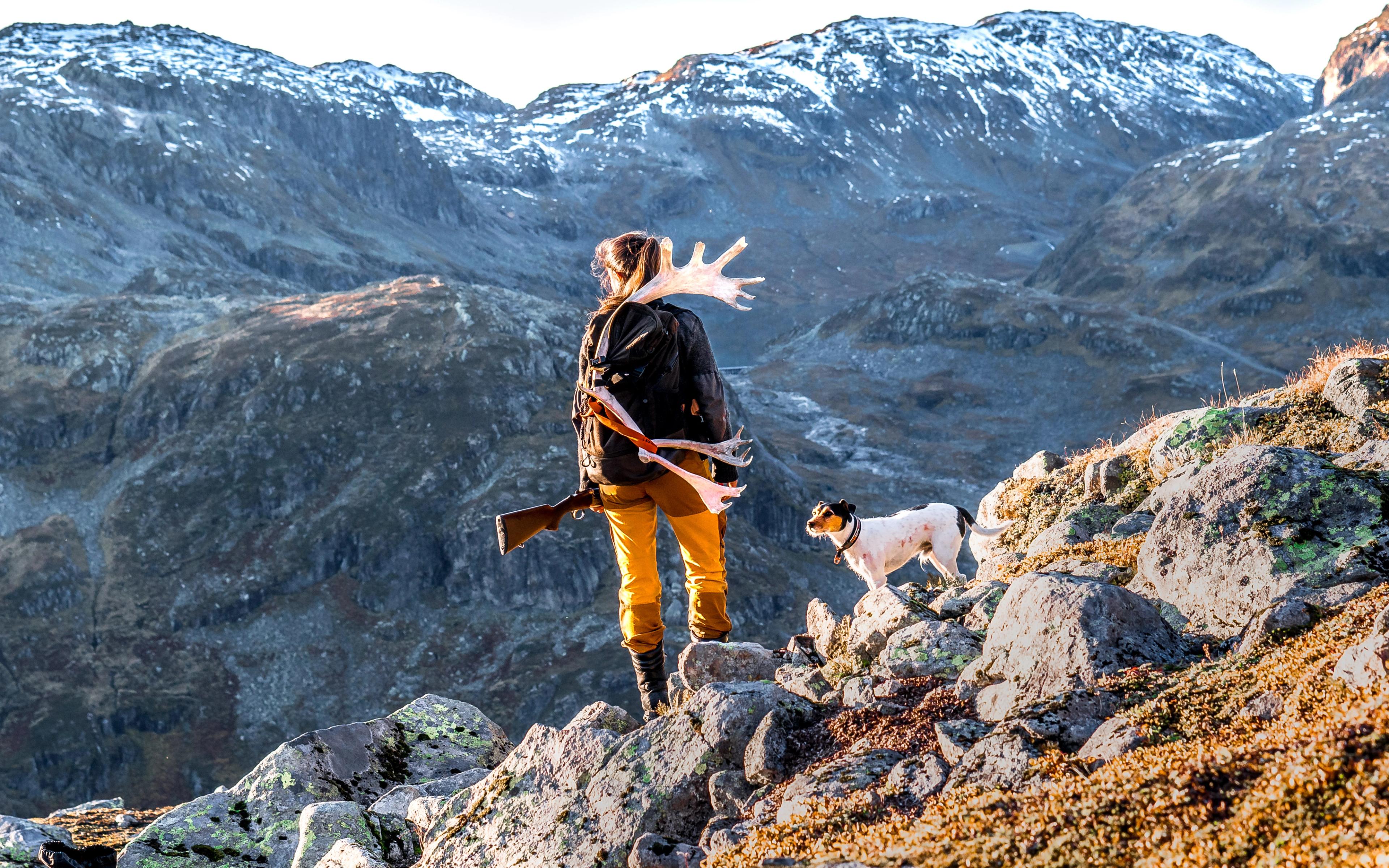 En kvinne står og ser utover fjellet med geværet i hånden og jakthunden ved sin side