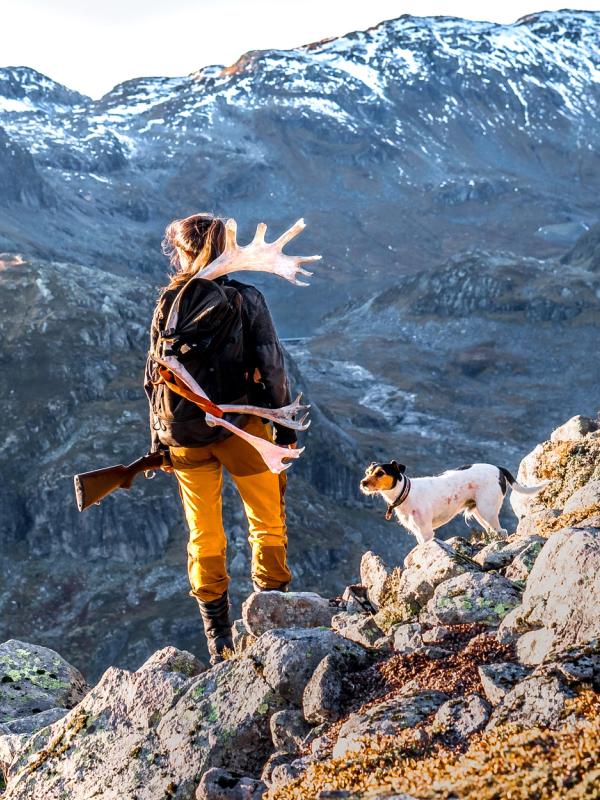 En kvinne står og ser utover fjellet med geværet i hånden og jakthunden ved sin side