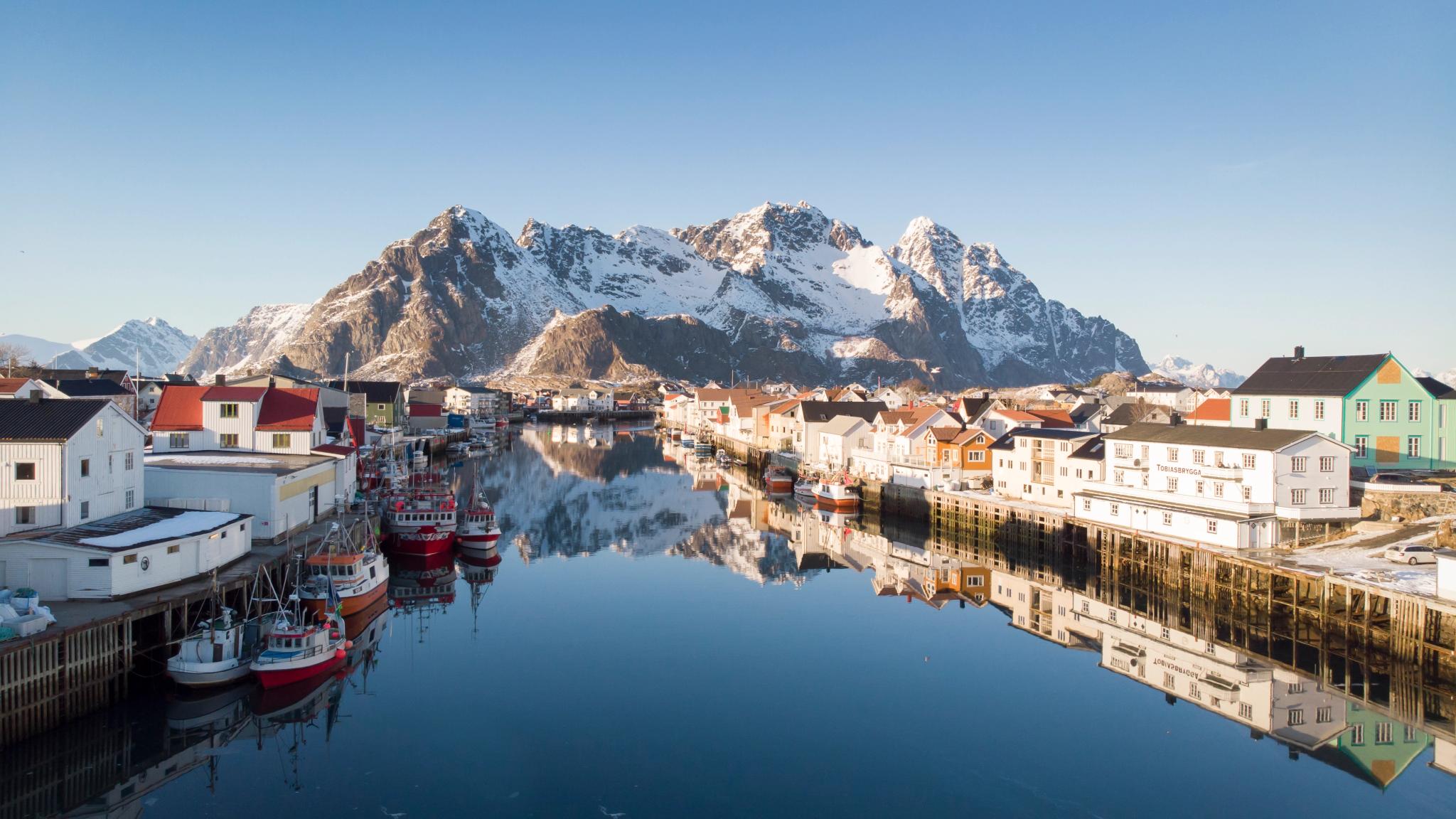 Trehus, fiskebåter og snødekte fjell i Henningsvær i Lofoten i Nord-Norge en solrik vinterdag