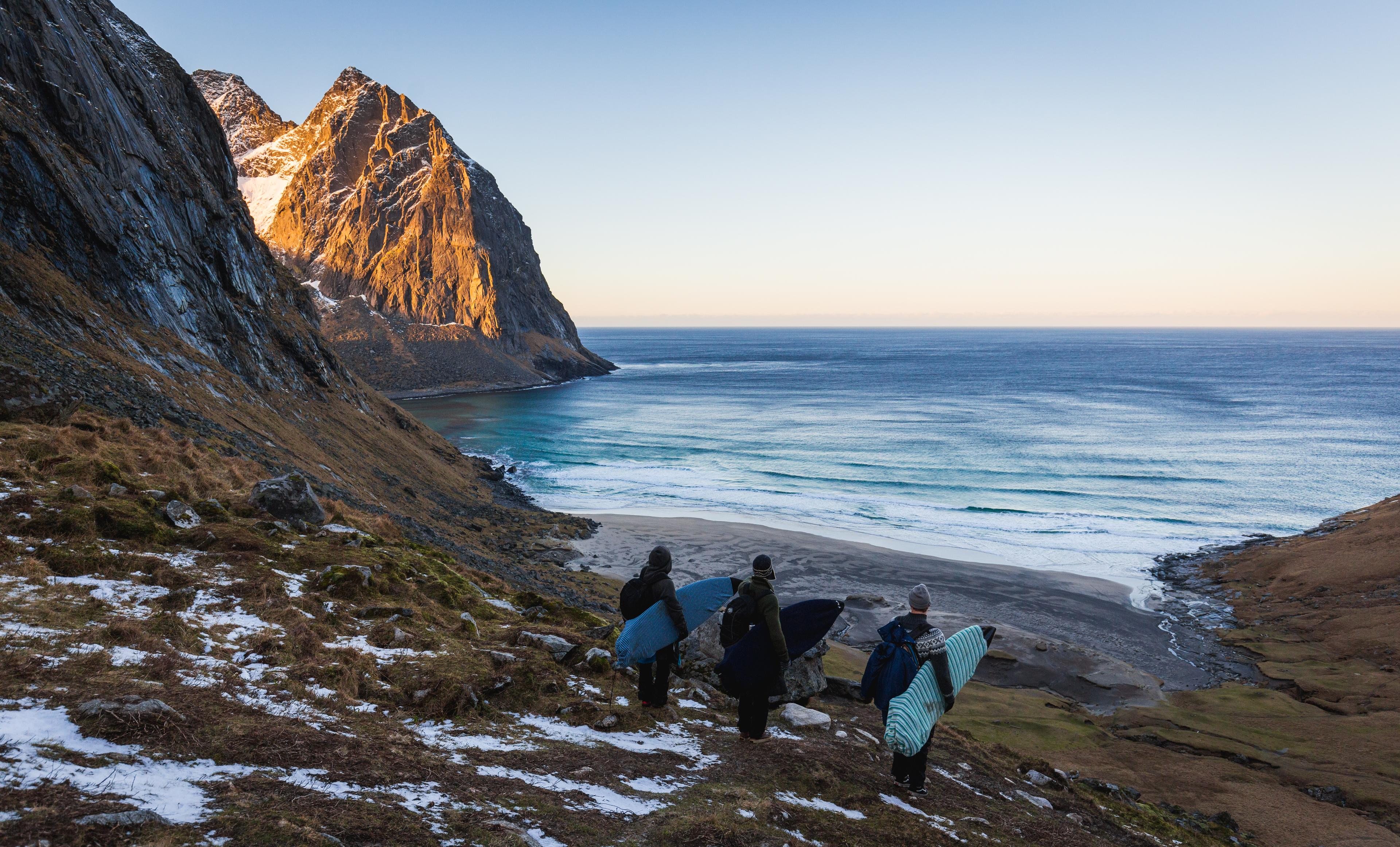 Three surfers with surf boards under their arms looking down at a bay in winter, Lofoten islands, Norhern Norway
