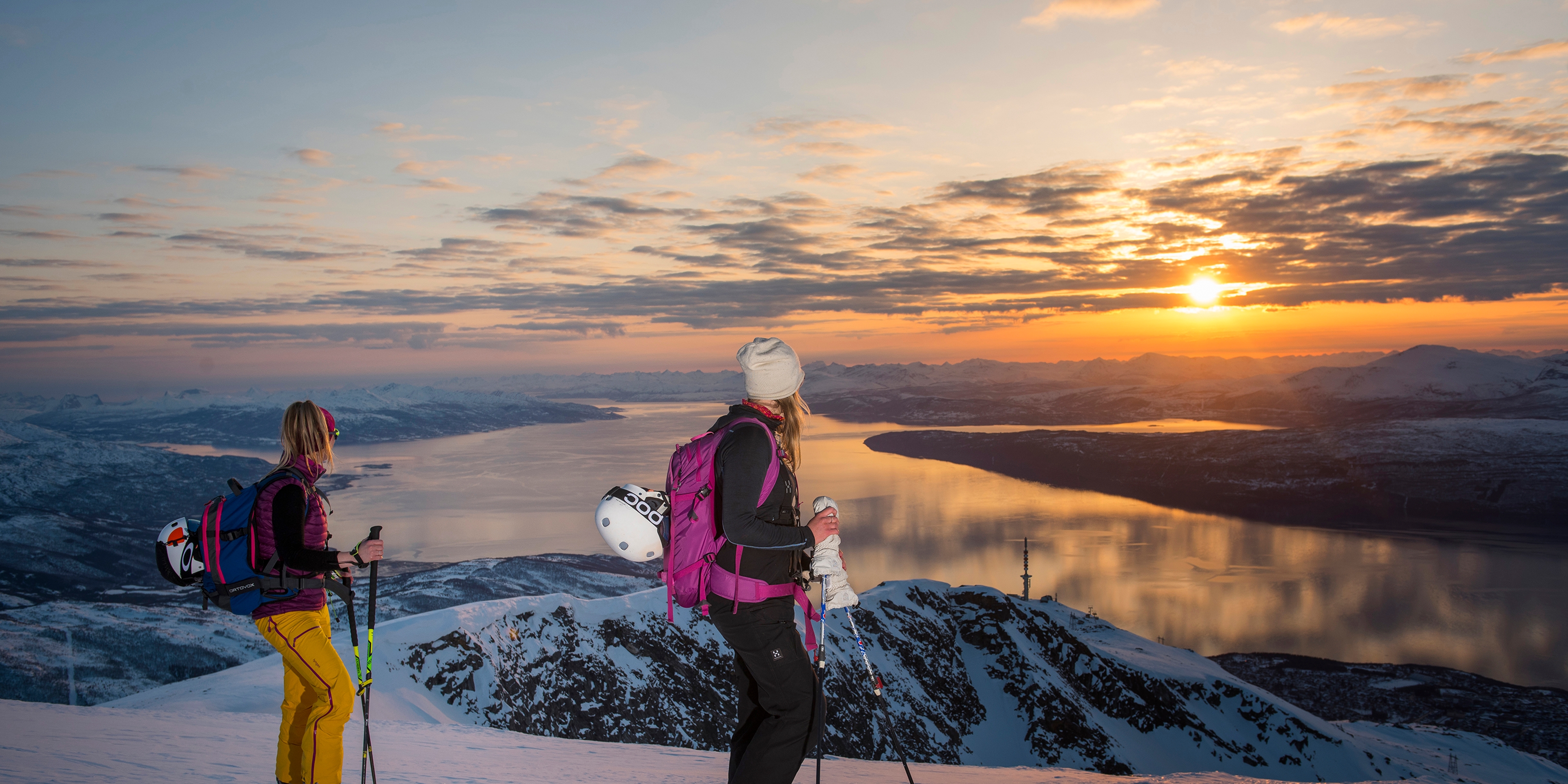 Two women with skis enjoying the view of mountains and fjord from the top of a mountain in Narvik, Northern Norway