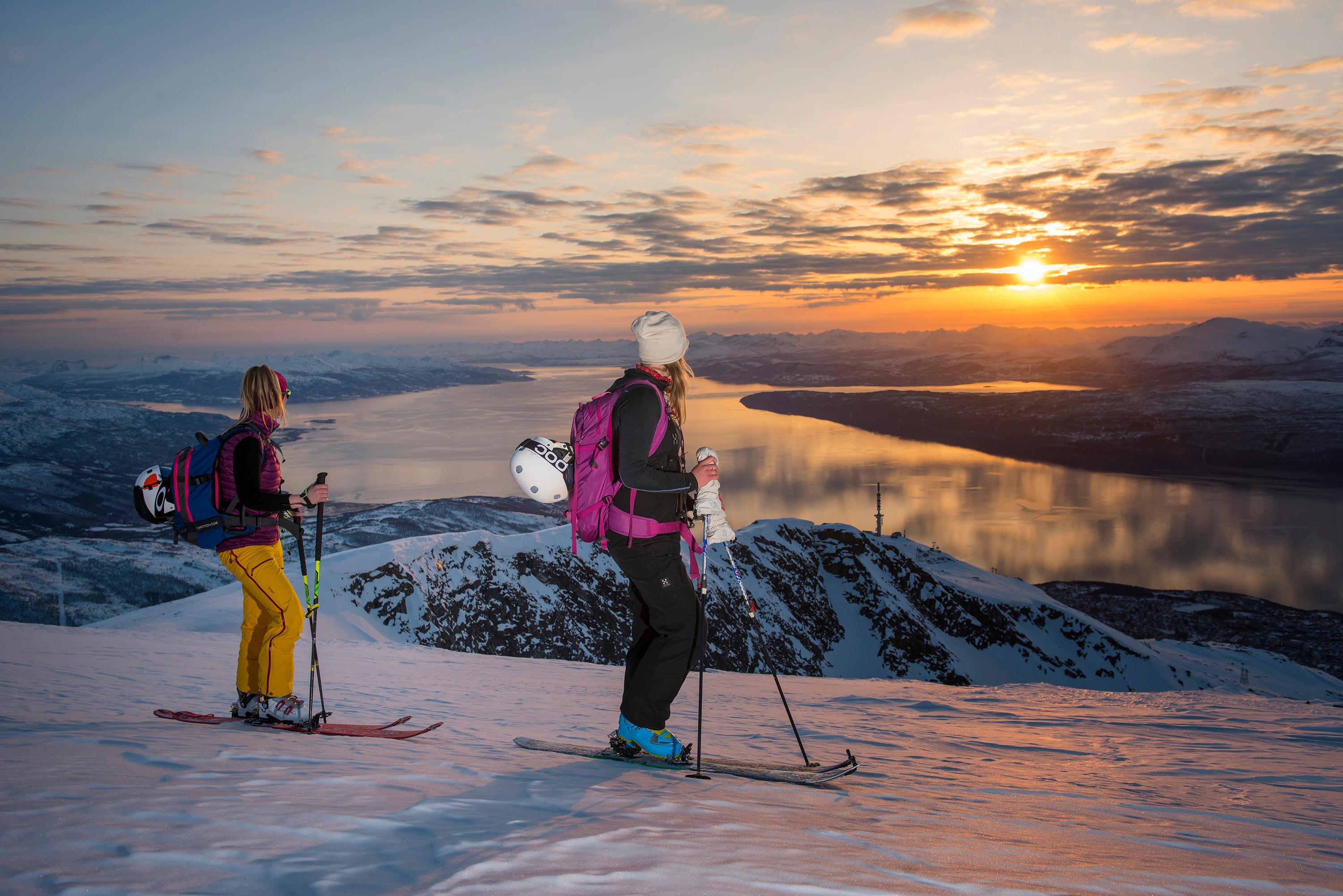 Two women with skis enjoying the view of mountains and fjord from the top of a mountain in Narvik, Northern Norway