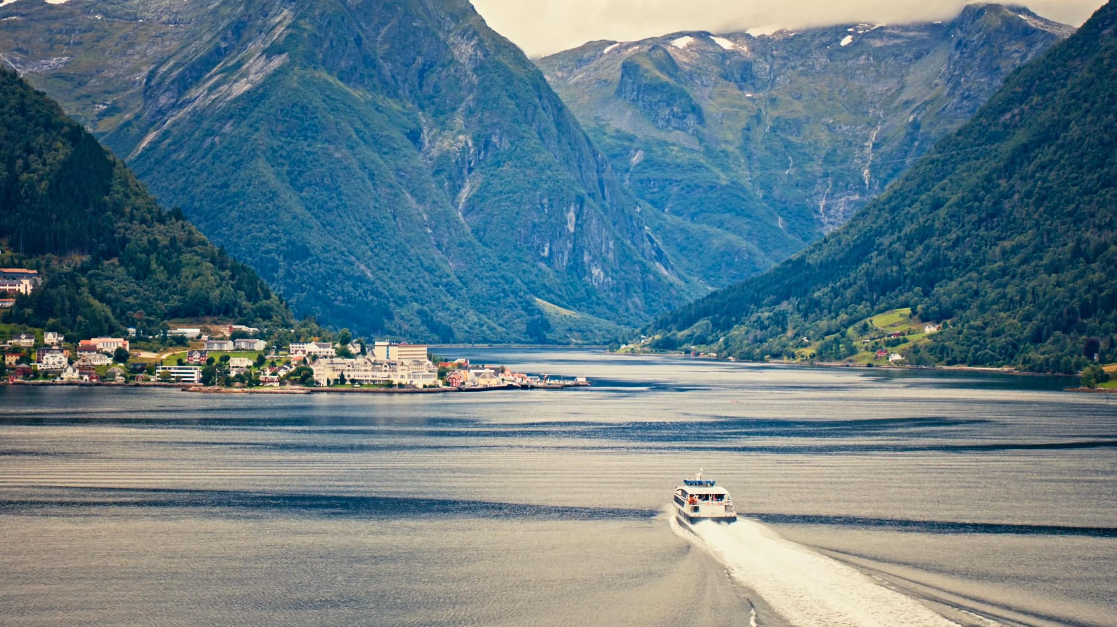 A boat sailing on a calm Norwegian fjord with a small village and mountains in the background
