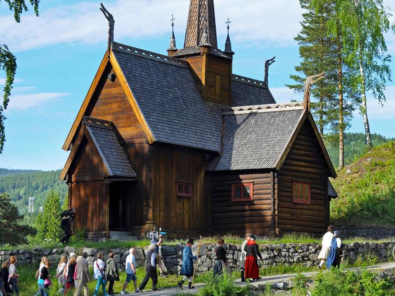 En gruppe mennesker i folkedragter går forbi stavkirken på Maihaugen i Lillehammer, Norge
