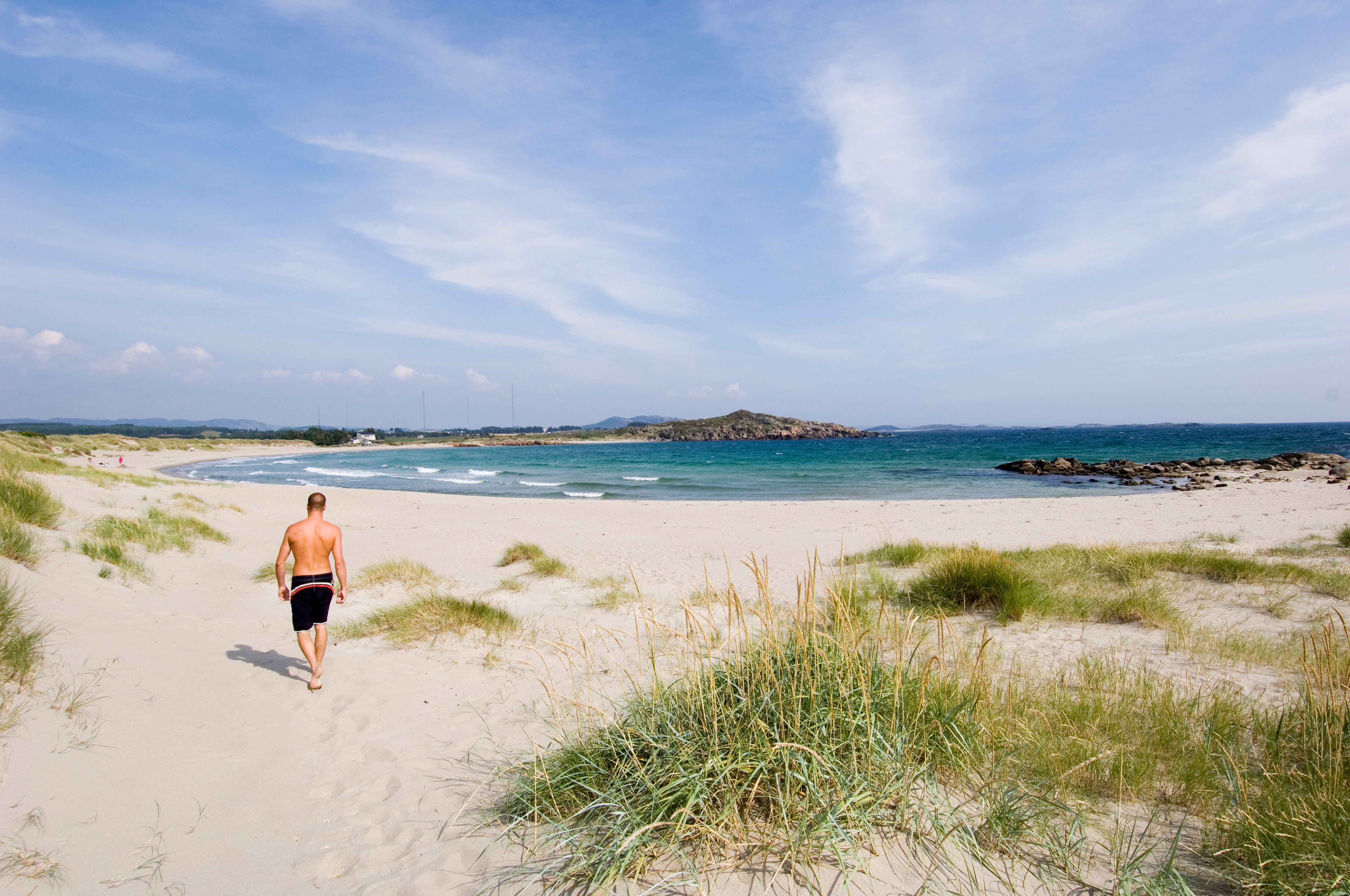 A man in a swim shorts walking on a white sandy beach in Lista, Southern Norway.