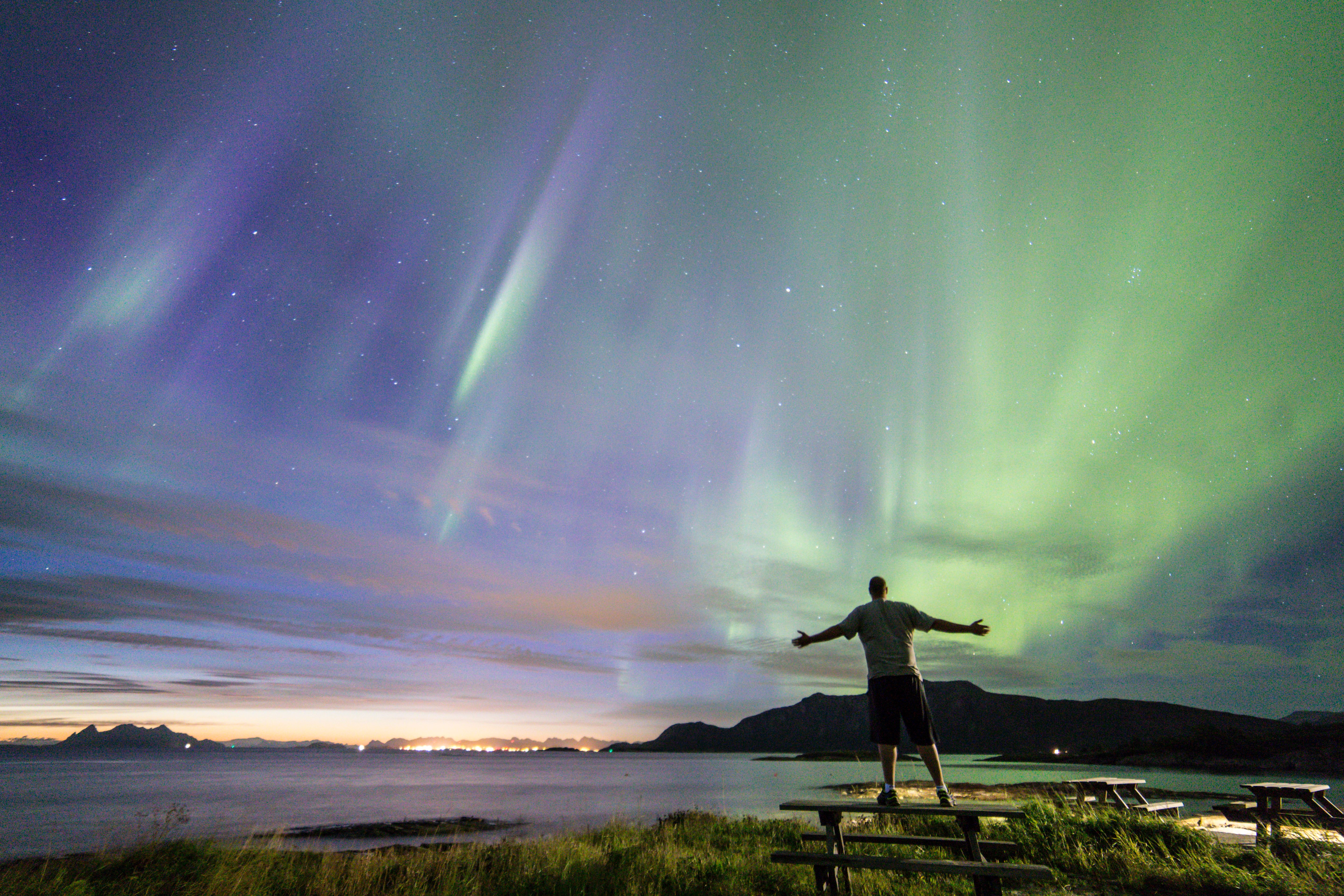 A person standing on a picknick table below green and violet northern lights at Gildeskål in Nordland, Northern Norway