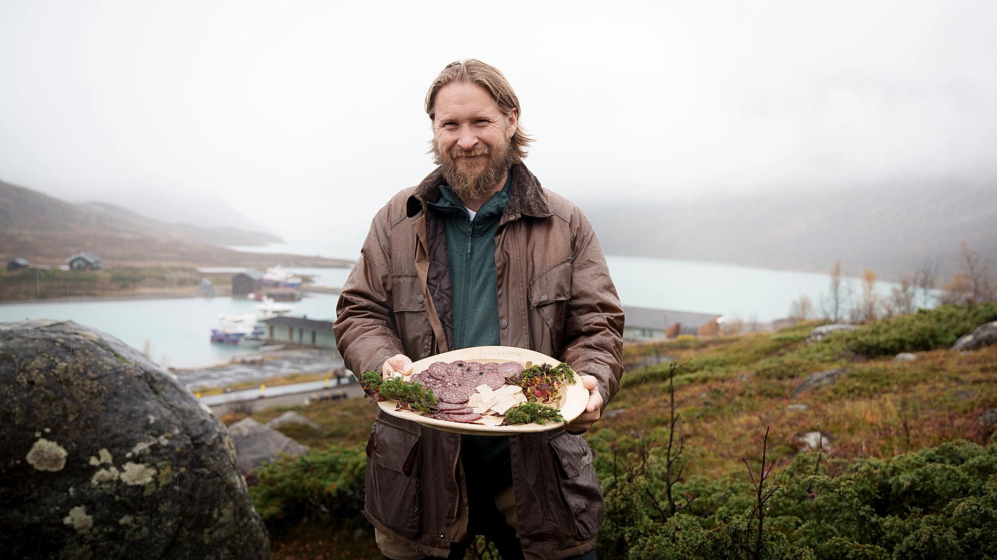 En mann i naturen holder et trefat med spekepølse.