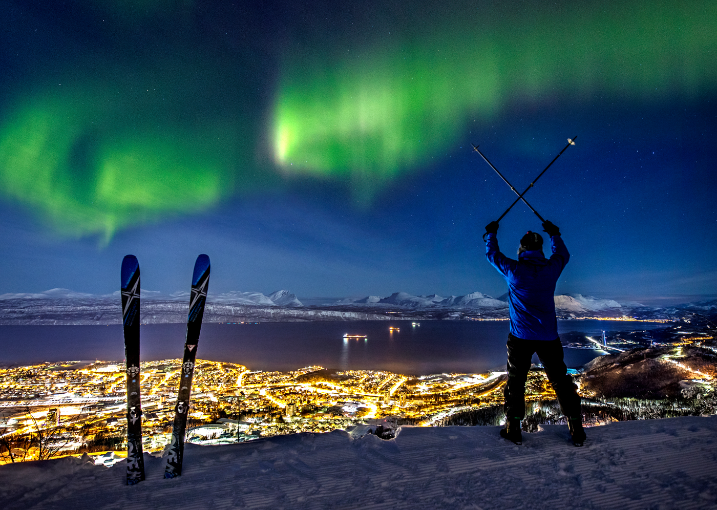 A man alpine skiing over Narvik town while spectacular northern lights
