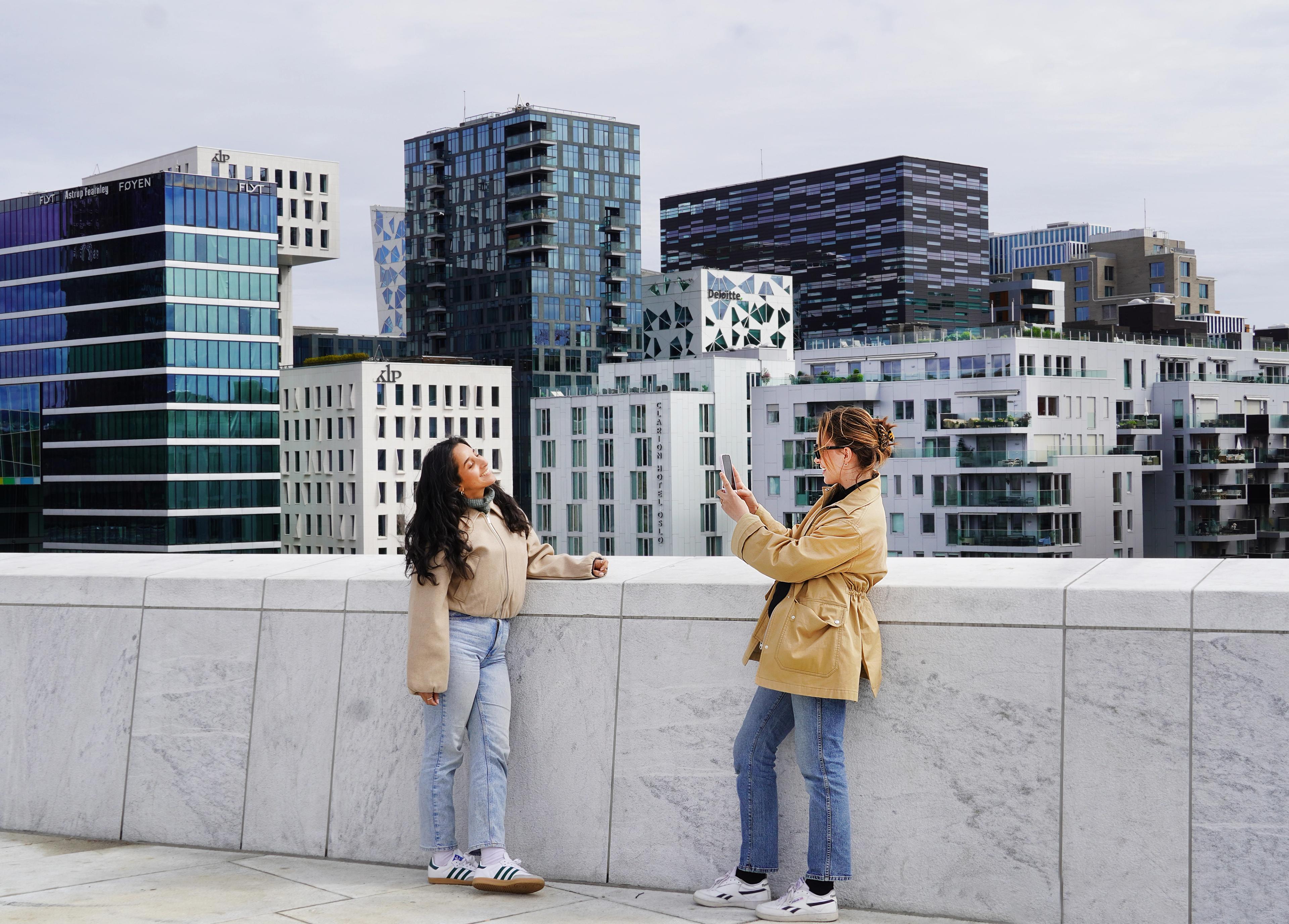 Two young women standing on the rooftop of the Oslo Opera House in Norway, capturing a photo with the city skyline in the background.