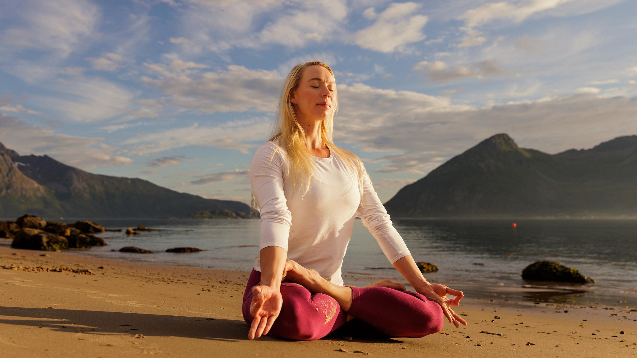 Ei dame sitter i lotusstilling å gjør yoga på stranden i midnattsolen i Nord-Norge