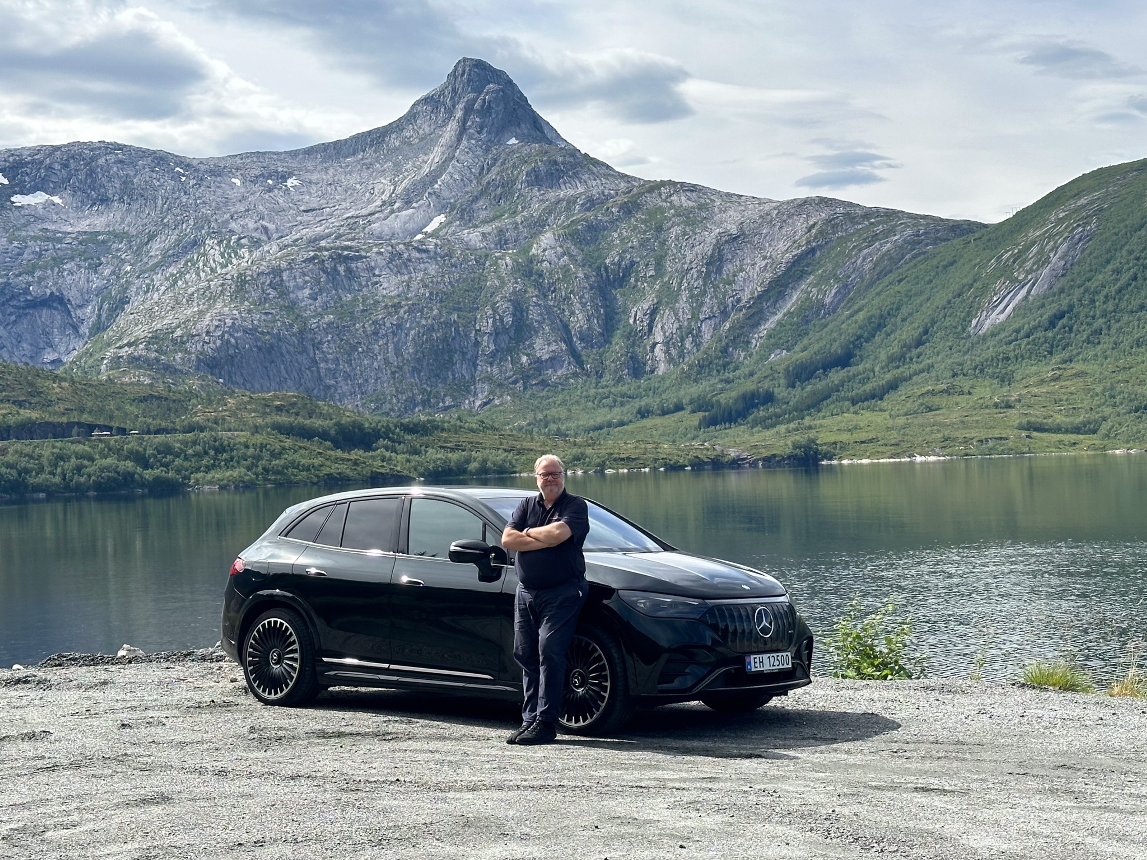 A man standing in front of an electric car with a body of water and a mountain behind him