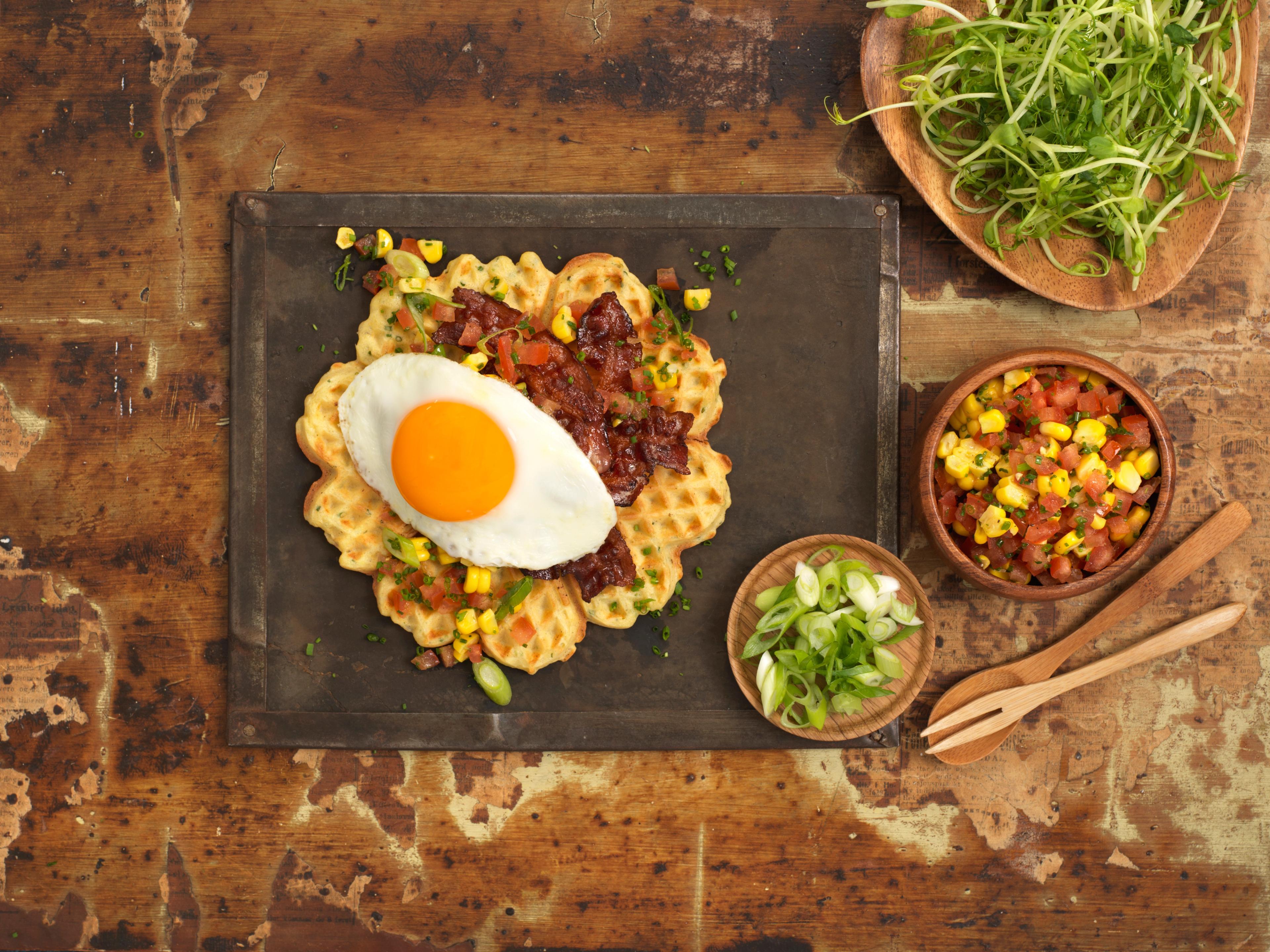 A plate with waffle with egg and bacon and some side dishes