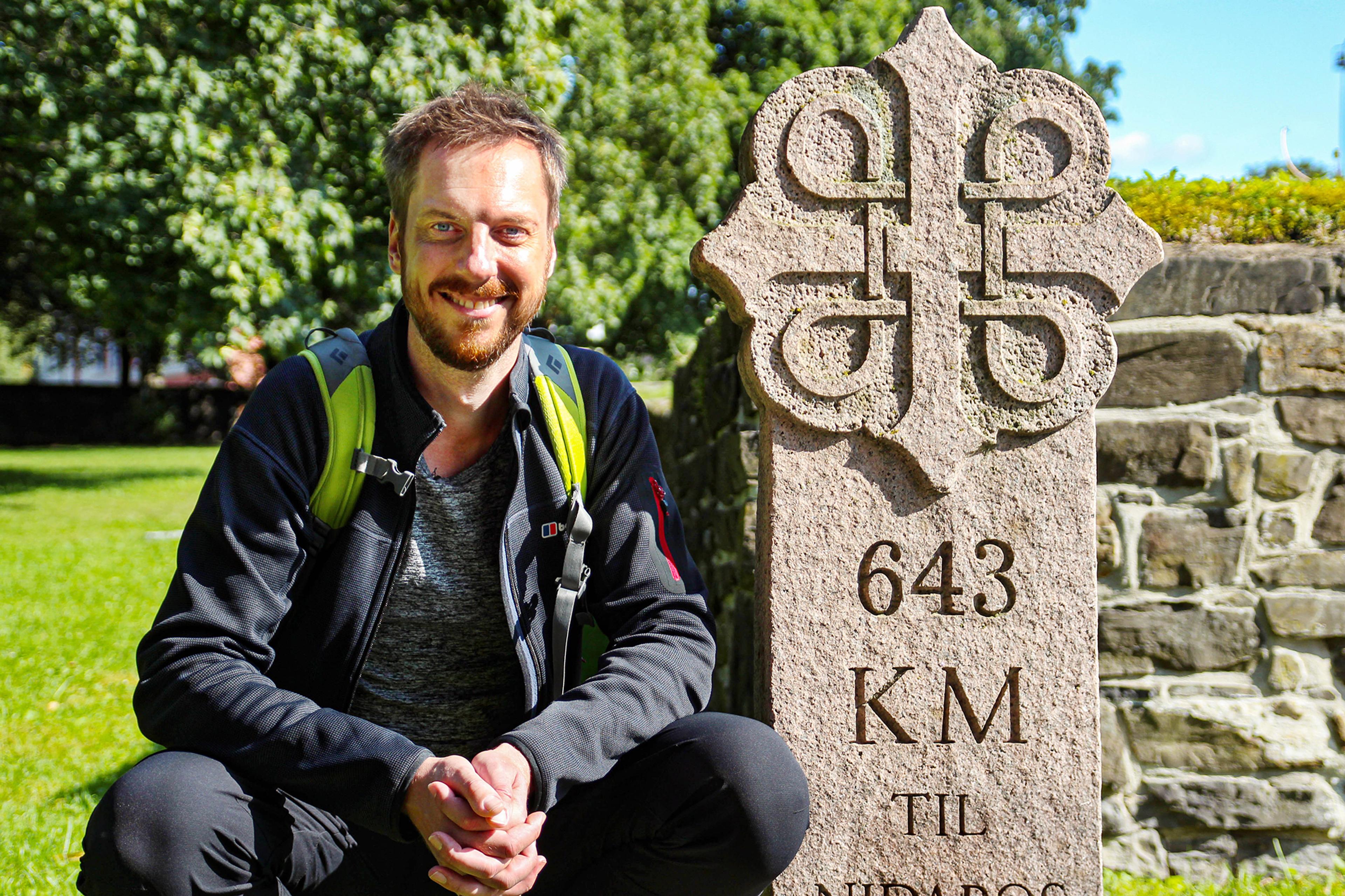 Pilgrimen Kes Blans bredvid en skylt som markerar början på pilgrimsvägen S:t Olavsleden från Oslo till Trondheim, Norge