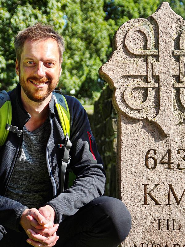 Pilgrimen Kes Blans bredvid en skylt som markerar början på pilgrimsvägen S:t Olavsleden från Oslo till Trondheim, Norge