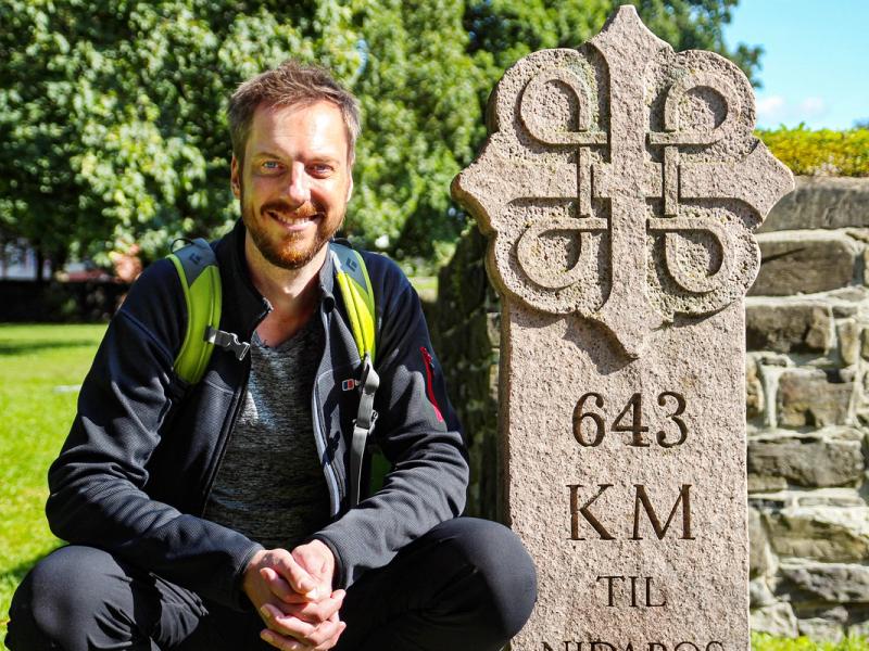 Pilgrimen Kes Blans bredvid en skylt som markerar början på pilgrimsvägen S:t Olavsleden från Oslo till Trondheim, Norge