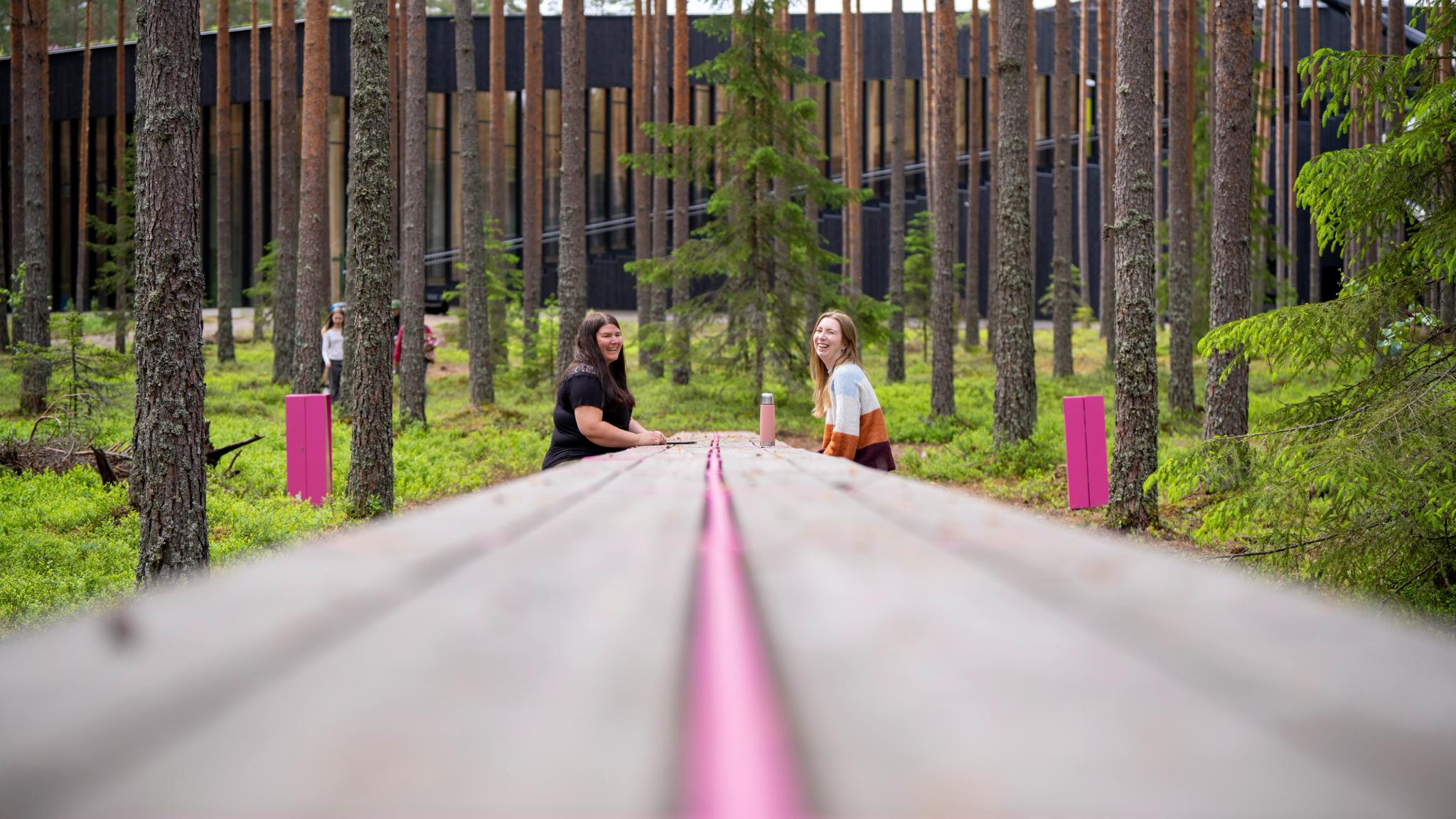 Two people sat at a long table in the forest next to The Plus furniture factory