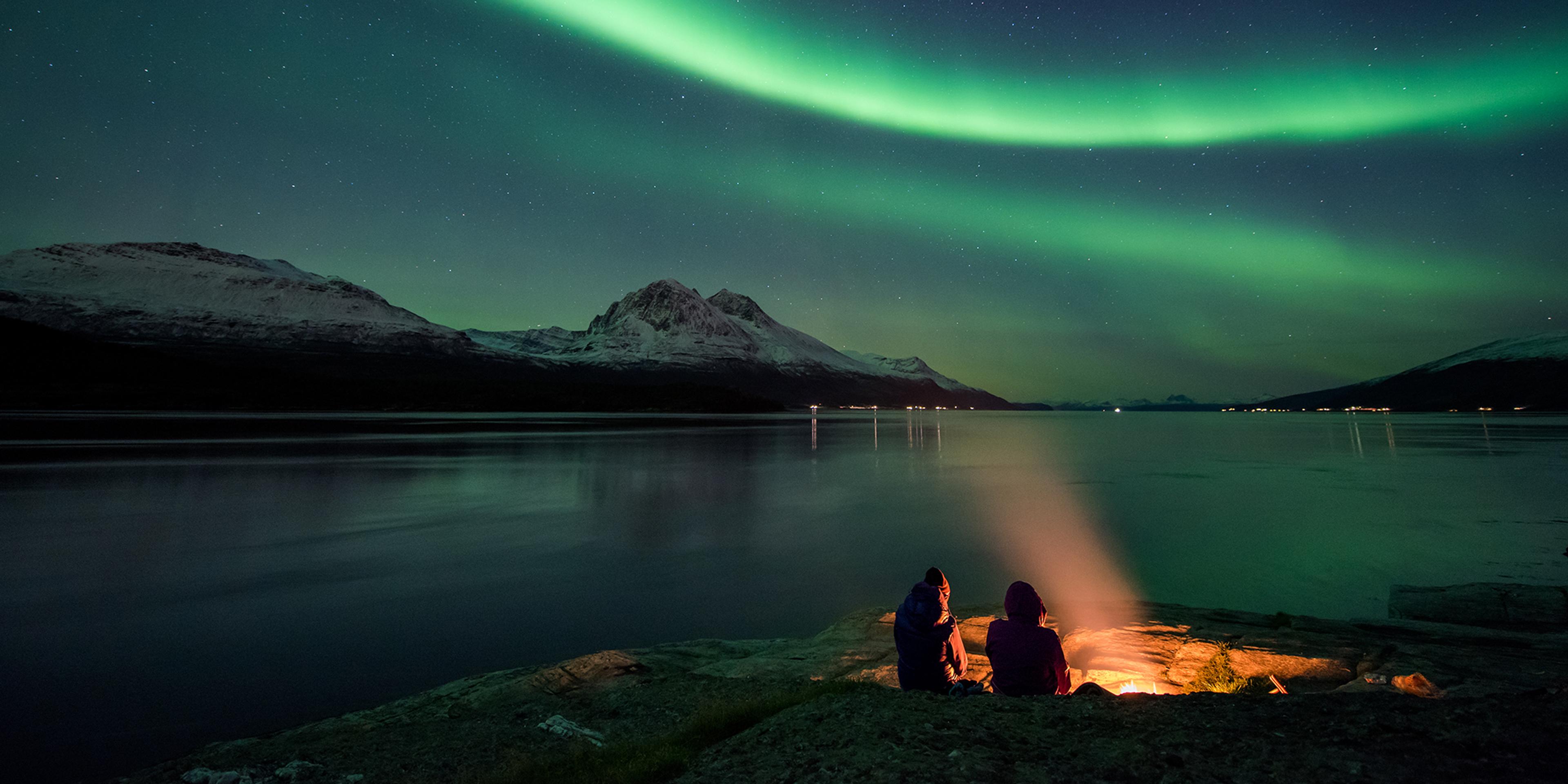 Two people sitting by a bonfire under the northern lights in Tromsø, Northern Norway