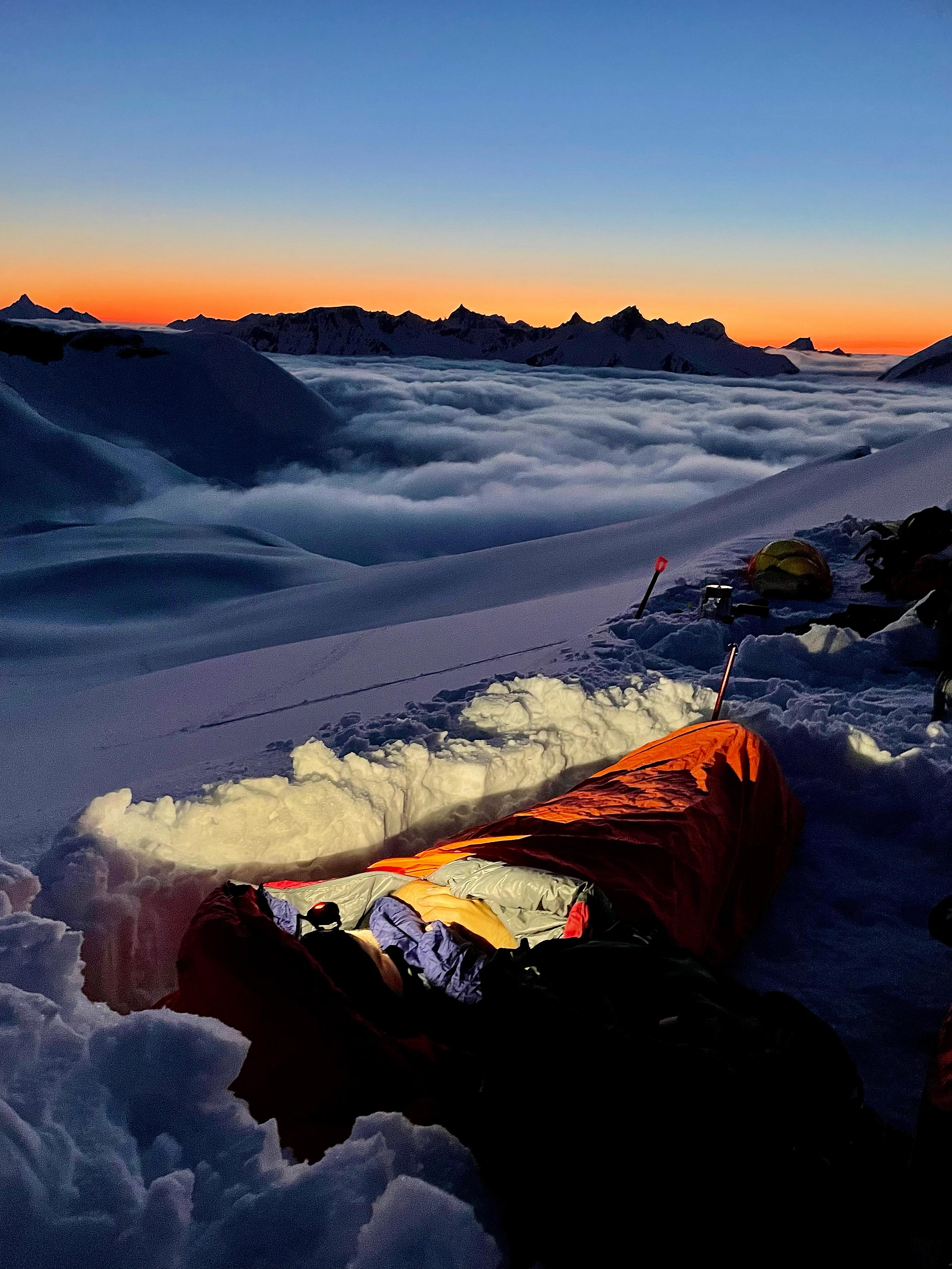 A man lying in a sleeping bag in the snow, enjoying the spectacular sunset of the Sunnmøre Alps in Ørsta, Fjord Norway