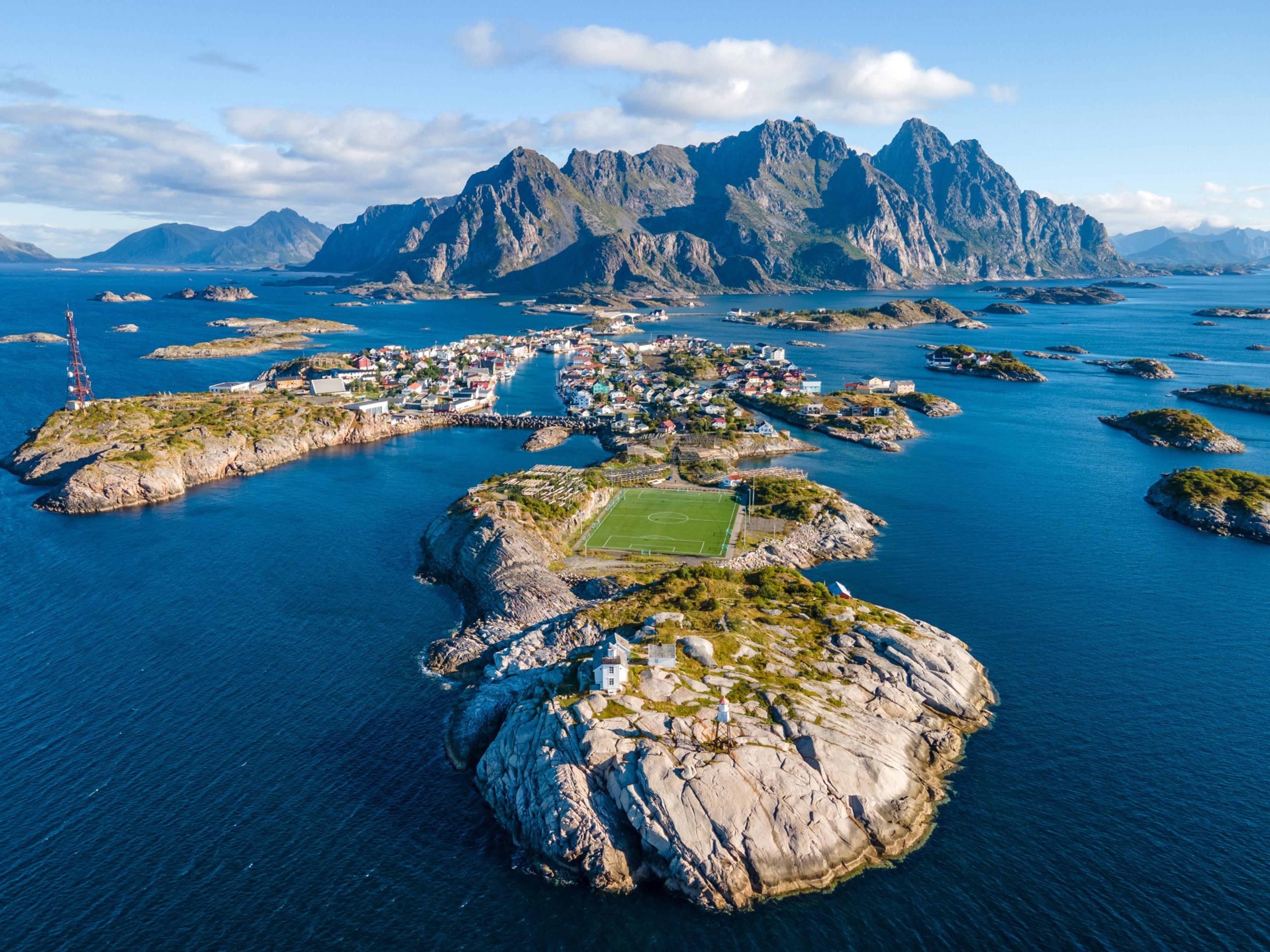 Coastal town on rocky islands with green football pitch facing mountain backdrop