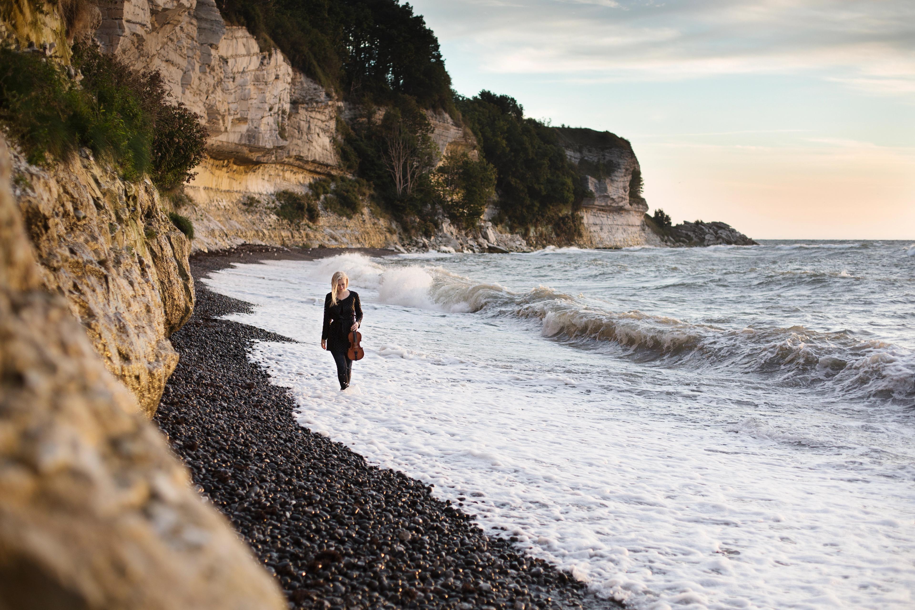 Eldbjørg Hemsing with her violin, walking along the coastline on a pebble beach, the waves touching her feet