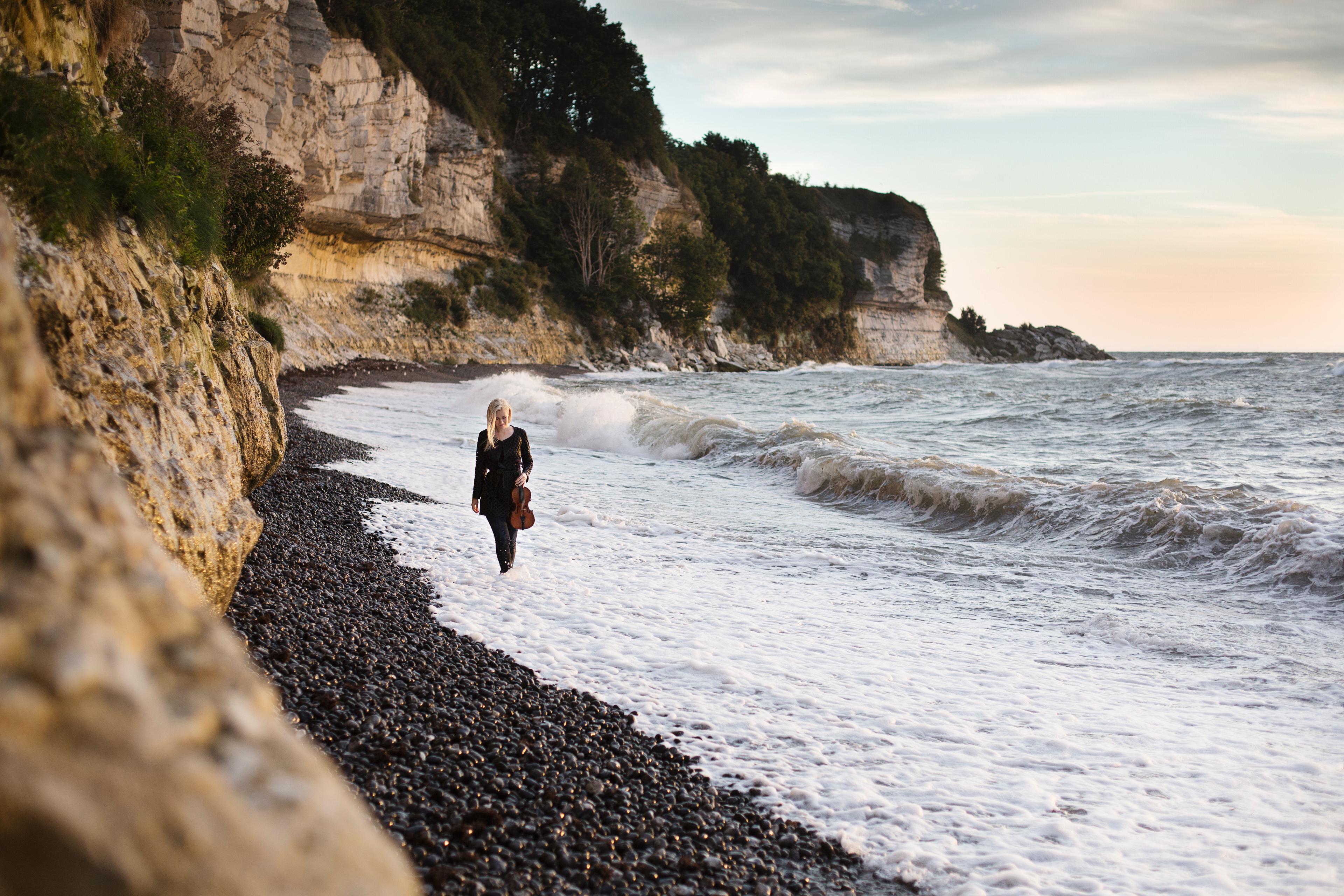 Eldbjørg Hemsing with her violin, walking along the coastline on a pebble beach, the waves touching her feet