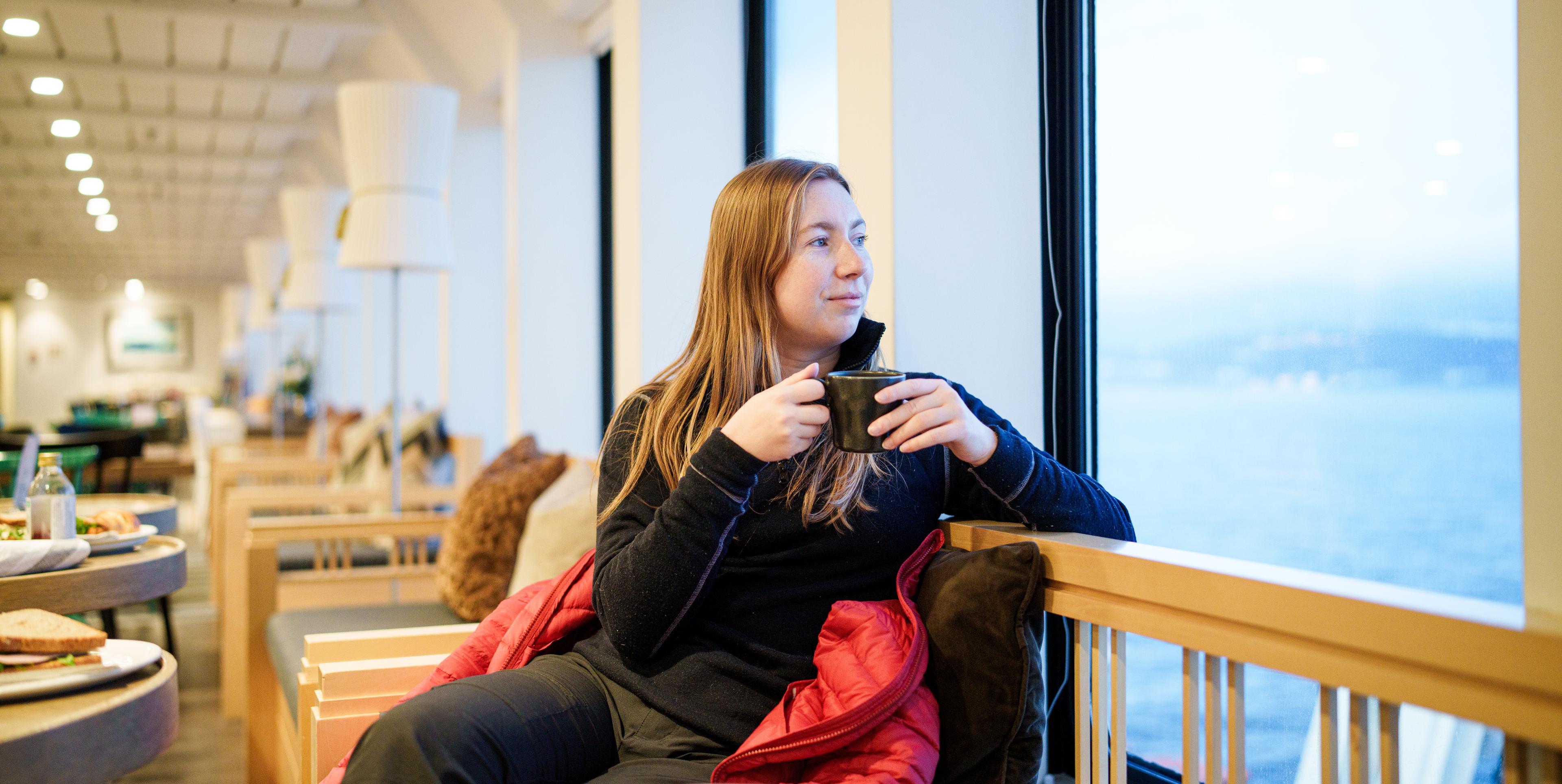 A woman holding a hot beverage and looking out at the view from the ship window