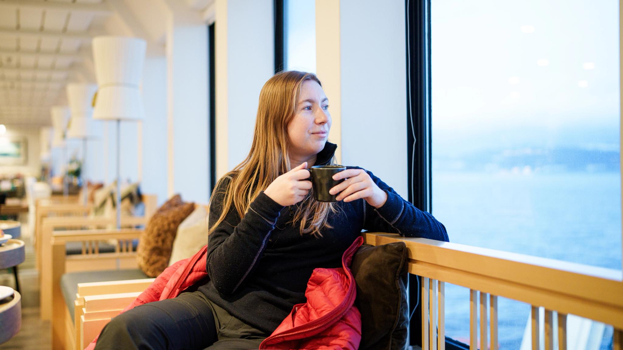 A woman holding a hot beverage and looking out at the view from the ship window