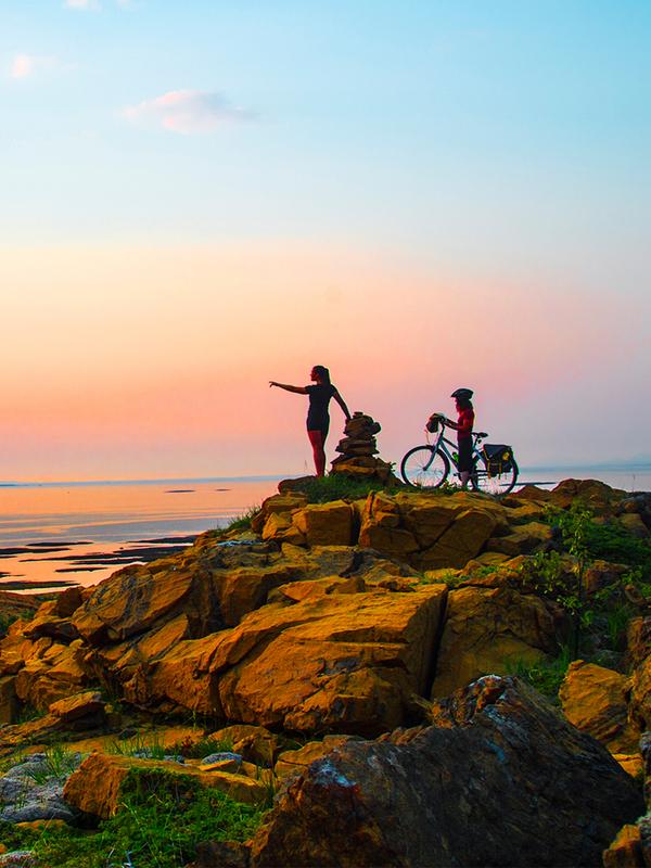 A hiker and a cyclist standing side by side watching the sunset at the Leka island in Trøndelag.