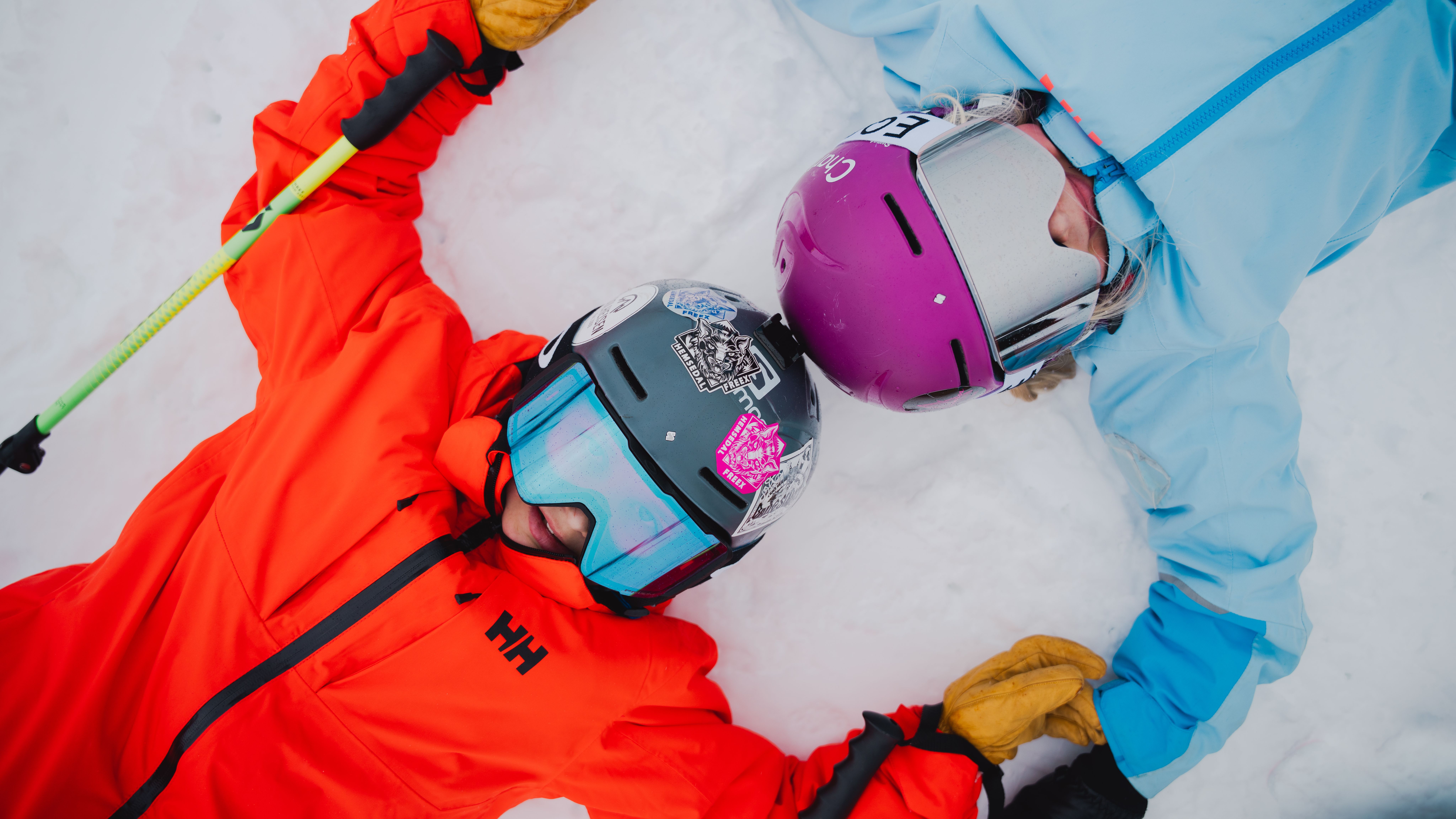 Two young kids in full alpine skiing gear, lying in the snow