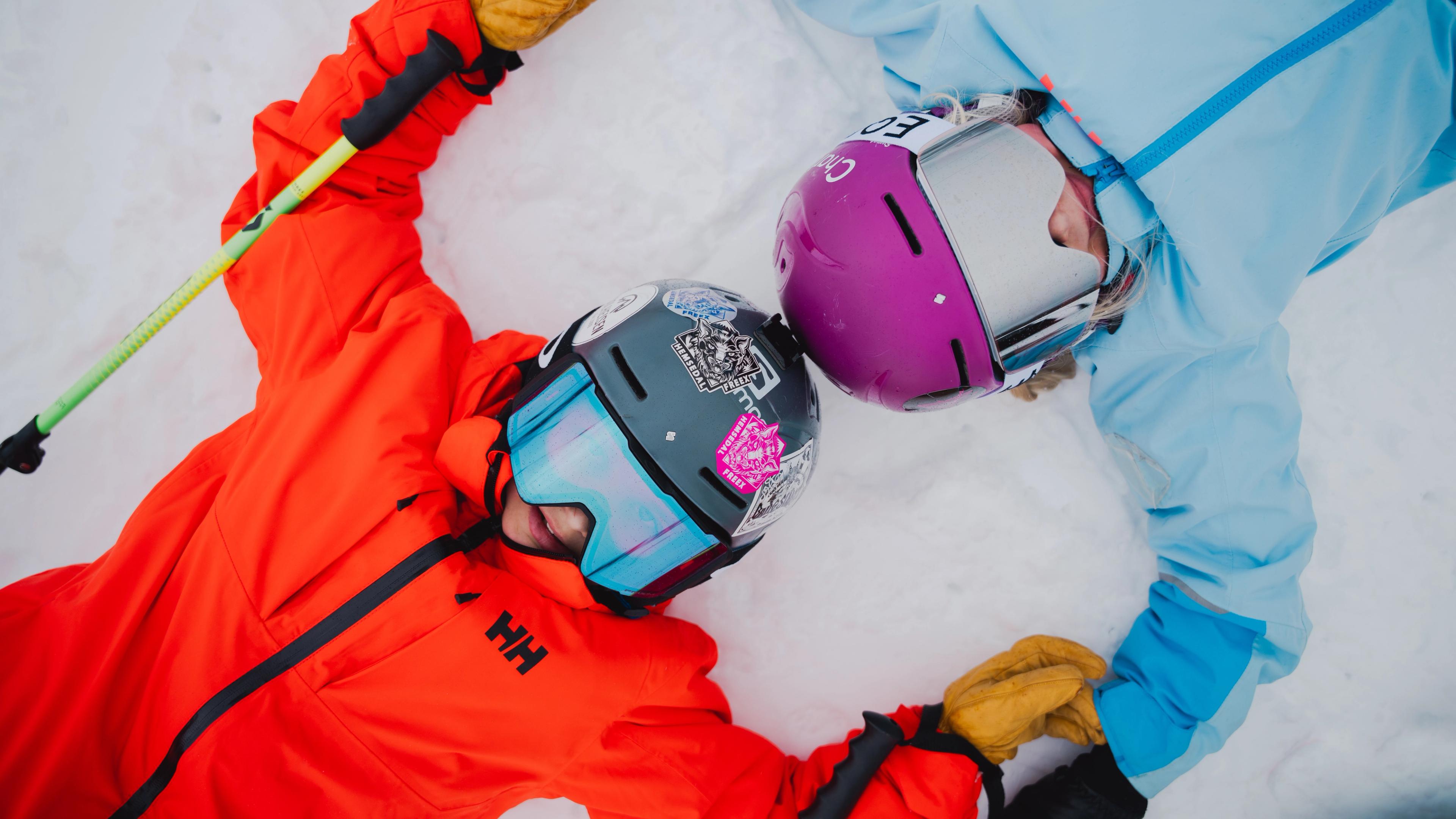 Two young kids in full alpine skiing gear, lying in the snow