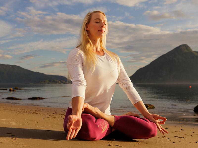 Ei dame sitter i lotusstilling å gjør yoga på stranden i midnattsolen i Nord-Norge