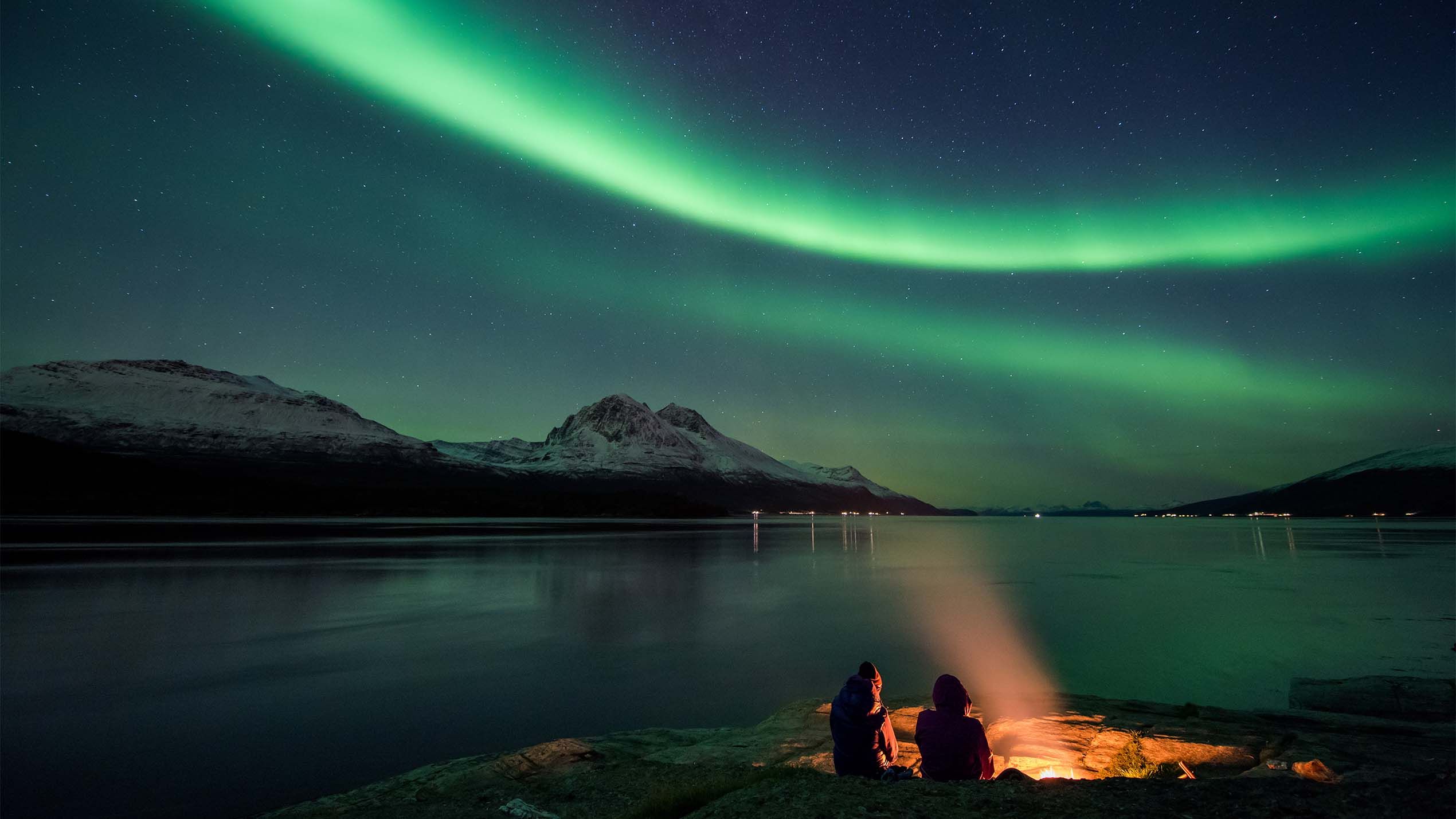 Two people are sitting next to a bonfire by the fjord in Tromsø, Northern Norway, admiring the northern lights.