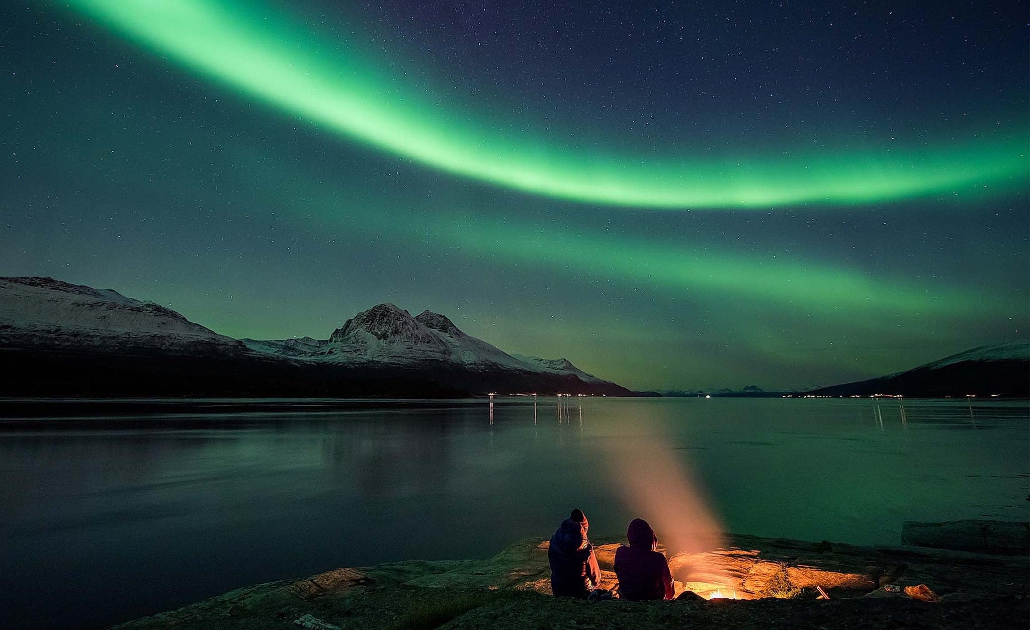 Two people are sitting next to a bonfire by the fjord in Tromsø, Northern Norway, admiring the northern lights.