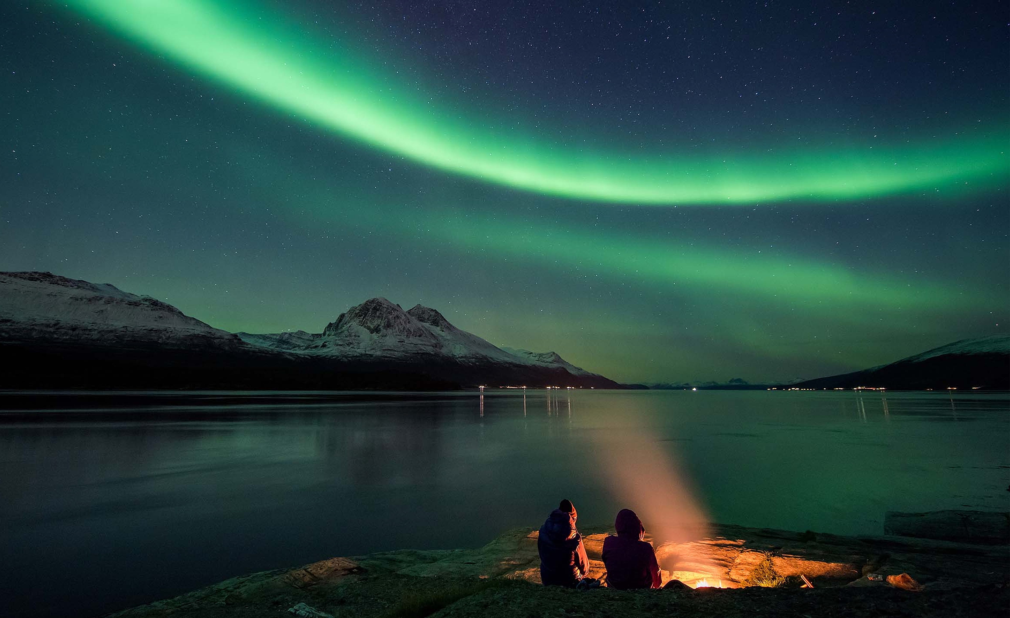 Two people are sitting next to a bonfire by the fjord in Tromsø, Northern Norway, admiring the northern lights.
