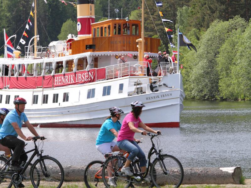 Un grupo de personas avanza en bicicleta por la ruta ciclista nacional que discurre junto al canal de Telemark, en el Este de Noruega, con el barco Henrik Ibsen al fondo.