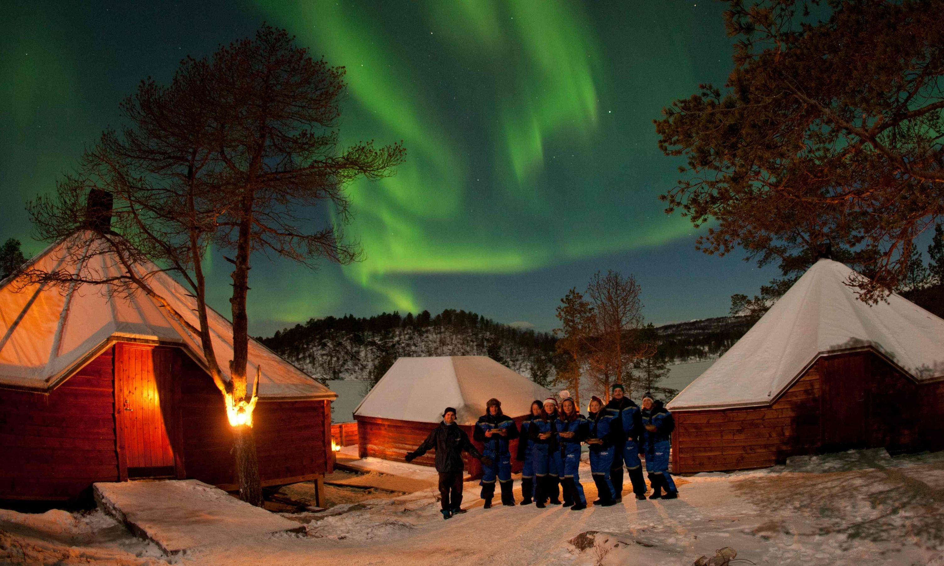 A group of people standing in front of several cabins under the northern lights