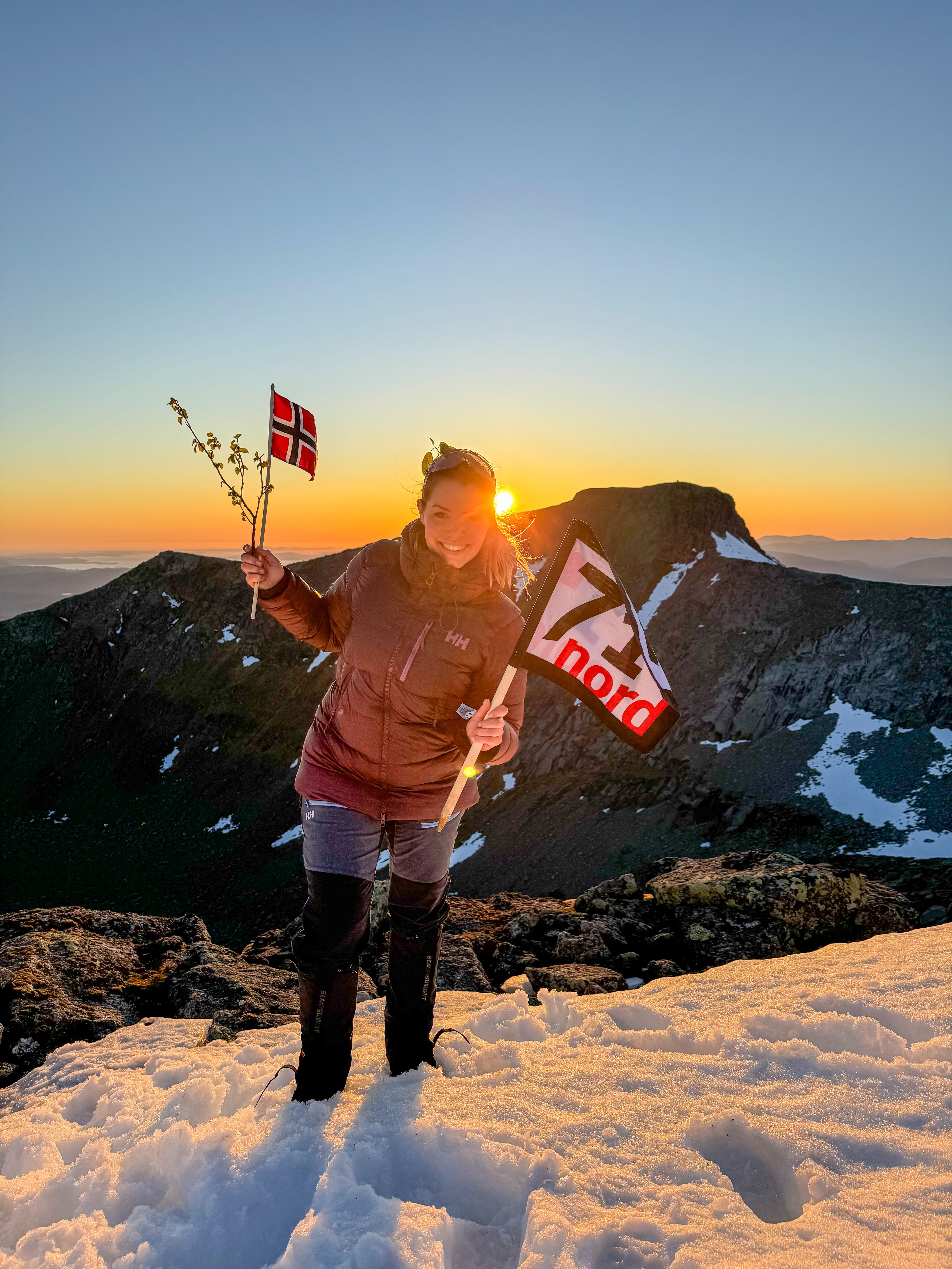 Woman waving the norwegian flag and a flag from the TV-production 71 grader nord in the sunset at a mountain in Rosendal, Hardanger