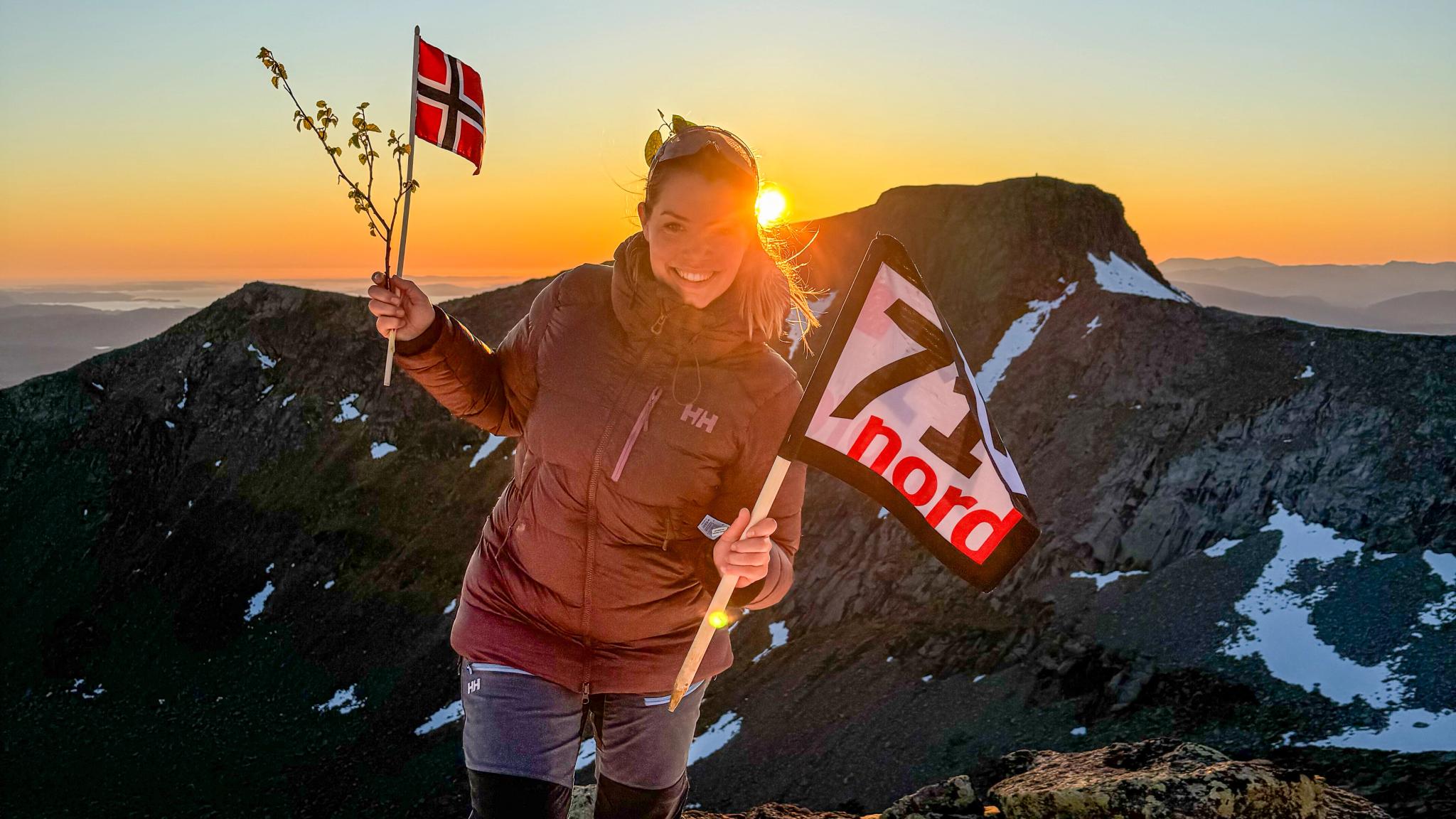 Woman waving the norwegian flag and a flag from the TV-production 71 grader nord in the sunset at a mountain in Rosendal, Hardanger