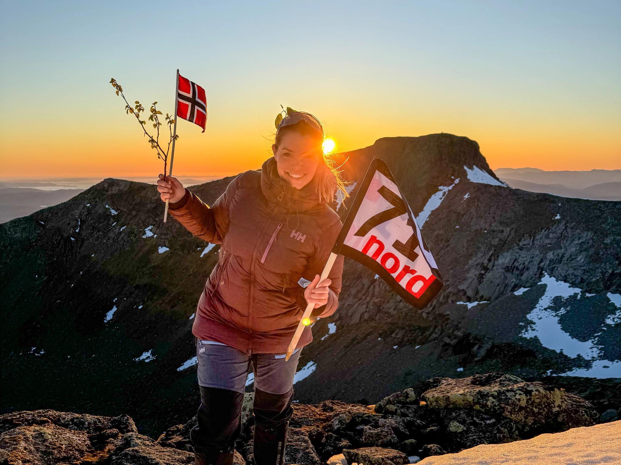 Woman waving the norwegian flag and a flag from the TV-production 71 grader nord in the sunset at a mountain in Rosendal, Hardanger