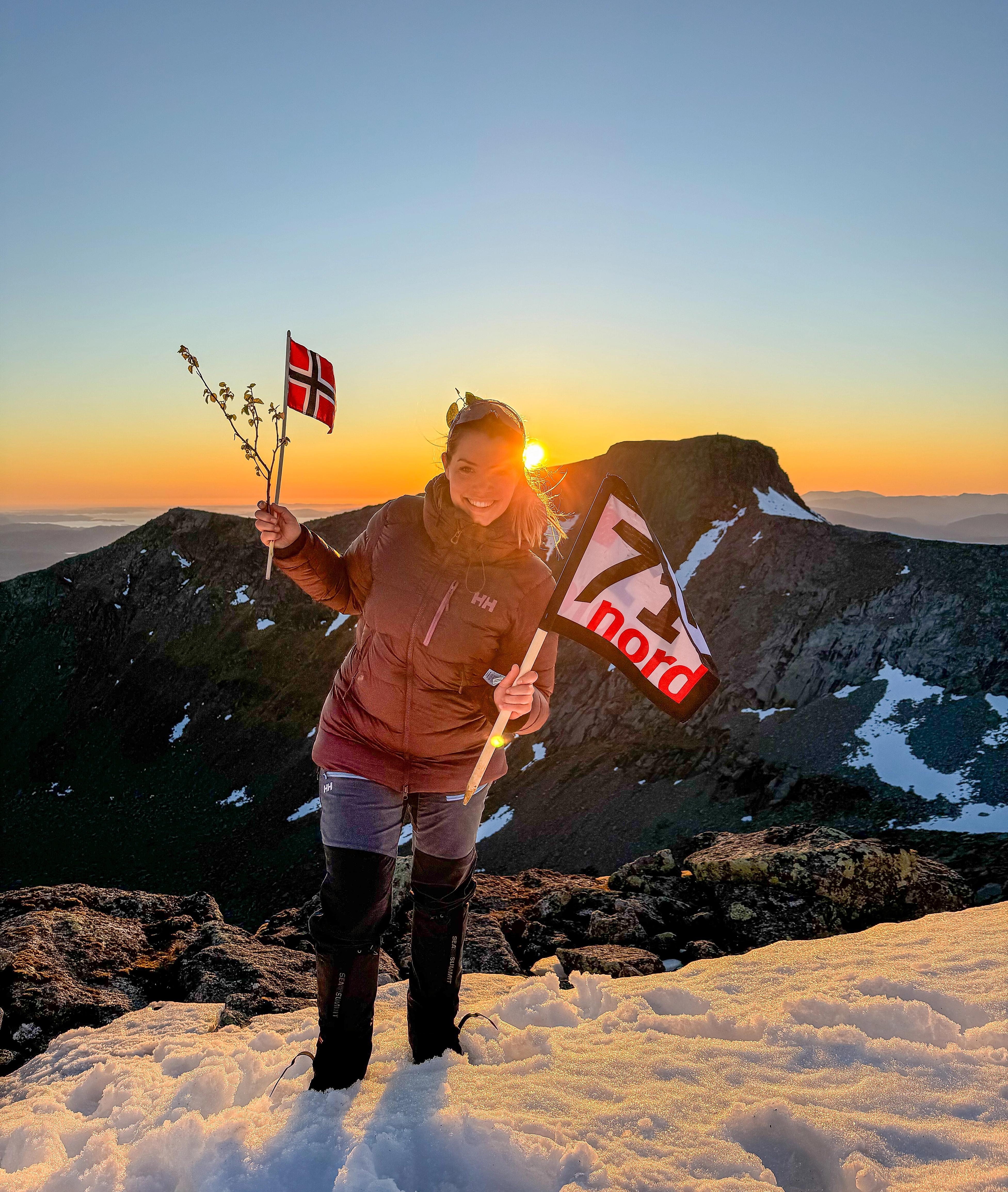 Woman waving the norwegian flag and a flag from the TV-production 71 grader nord in the sunset at a mountain in Rosendal, Hardanger