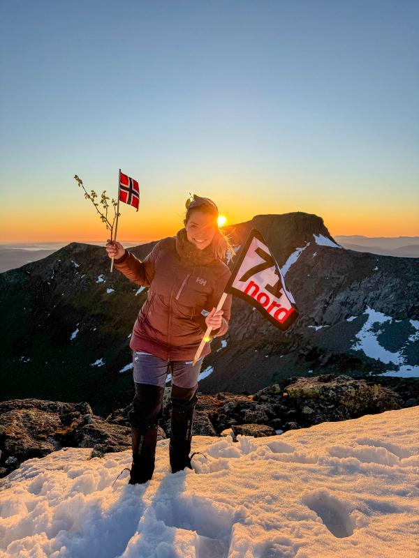 Woman waving the norwegian flag and a flag from the TV-production 71 grader nord in the sunset at a mountain in Rosendal, Hardanger