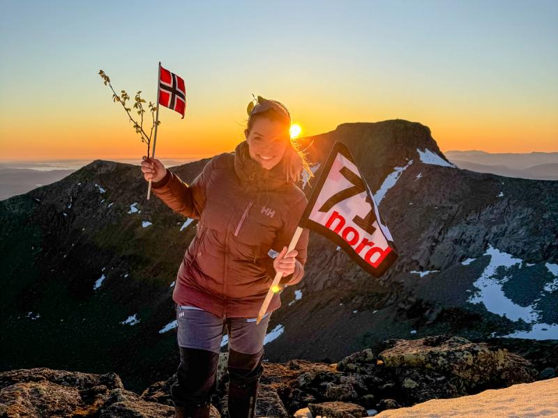 Woman waving the norwegian flag and a flag from the TV-production 71 grader nord in the sunset at a mountain in Rosendal, Hardanger