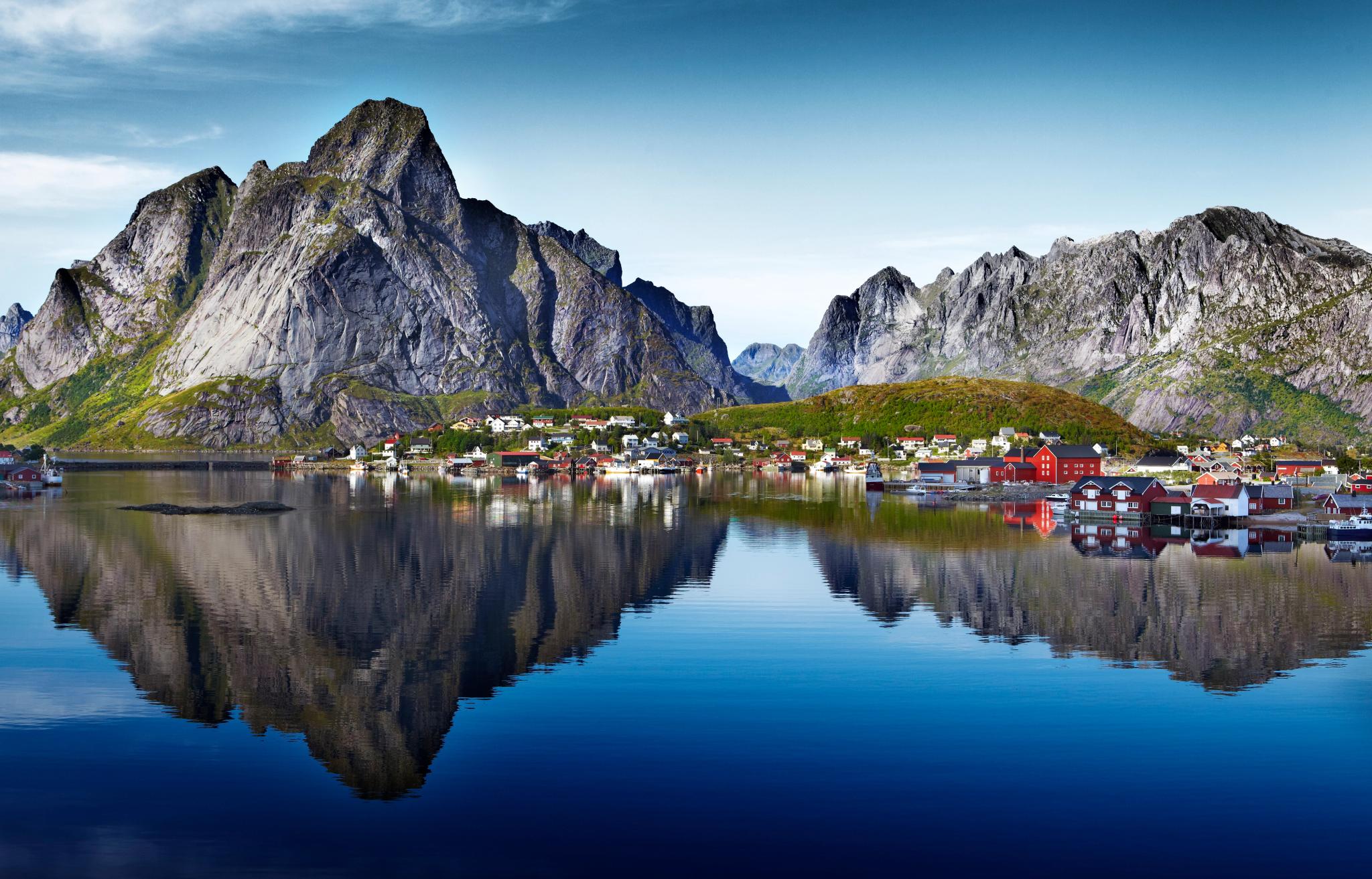 Fishing village in the Lofoten Islands, with red and white buildings reflected in calm water and dramatic mountain peaks behind.
