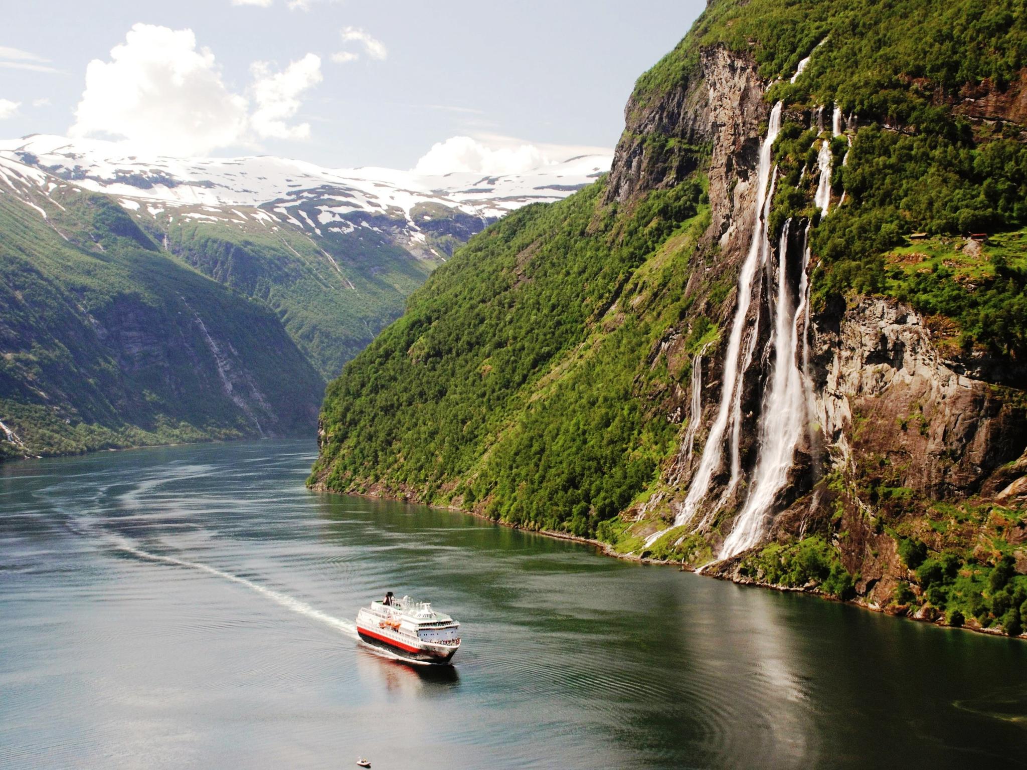 A Hurtigruten ship sailing through Geirangerfjord past the Seven Sisters waterfall, with snow-capped mountains in the background.