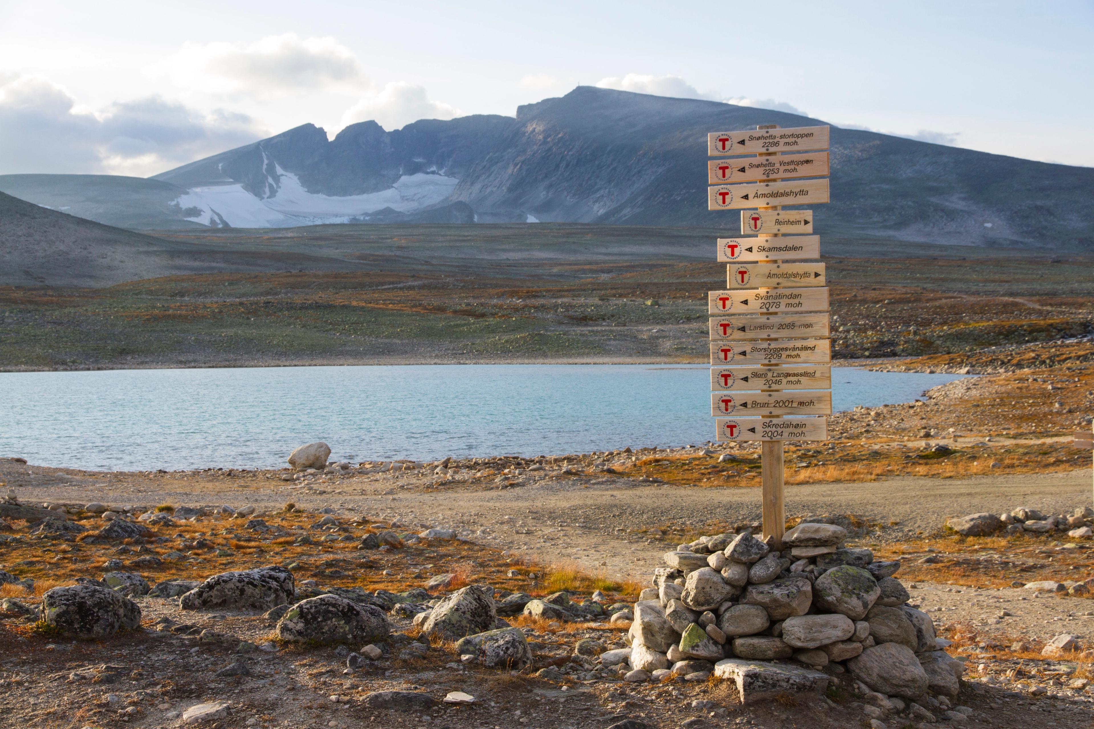 A small cairn holding a wooden pole with several signs with a lake and the Snøhetta mountain in the background. Dovrefjell, Eastern Norway.
