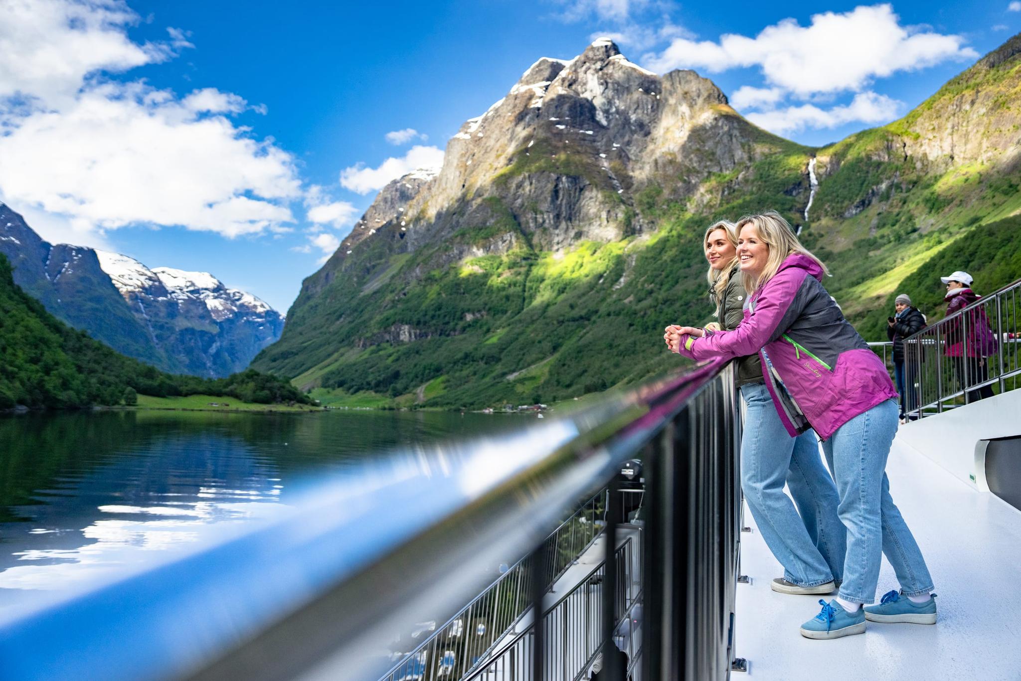 Two women viewing scenic Norwegian fjord from a boat, with dramatic mountains reflected in calm water under blue sky.